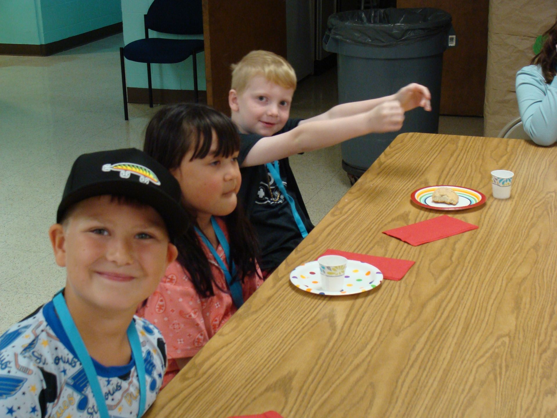 Three children sitting at a table with plates and cups, smiling and waving at the camera.