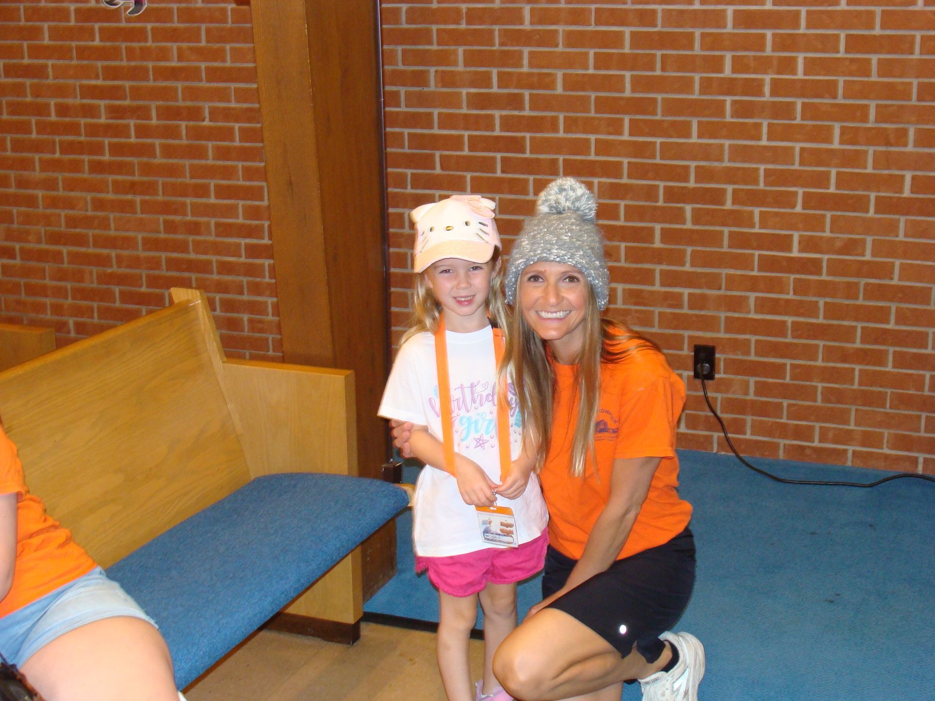 Two people kneel indoors beside a blue bench, smiling and wearing colorful hats against a brick wall.