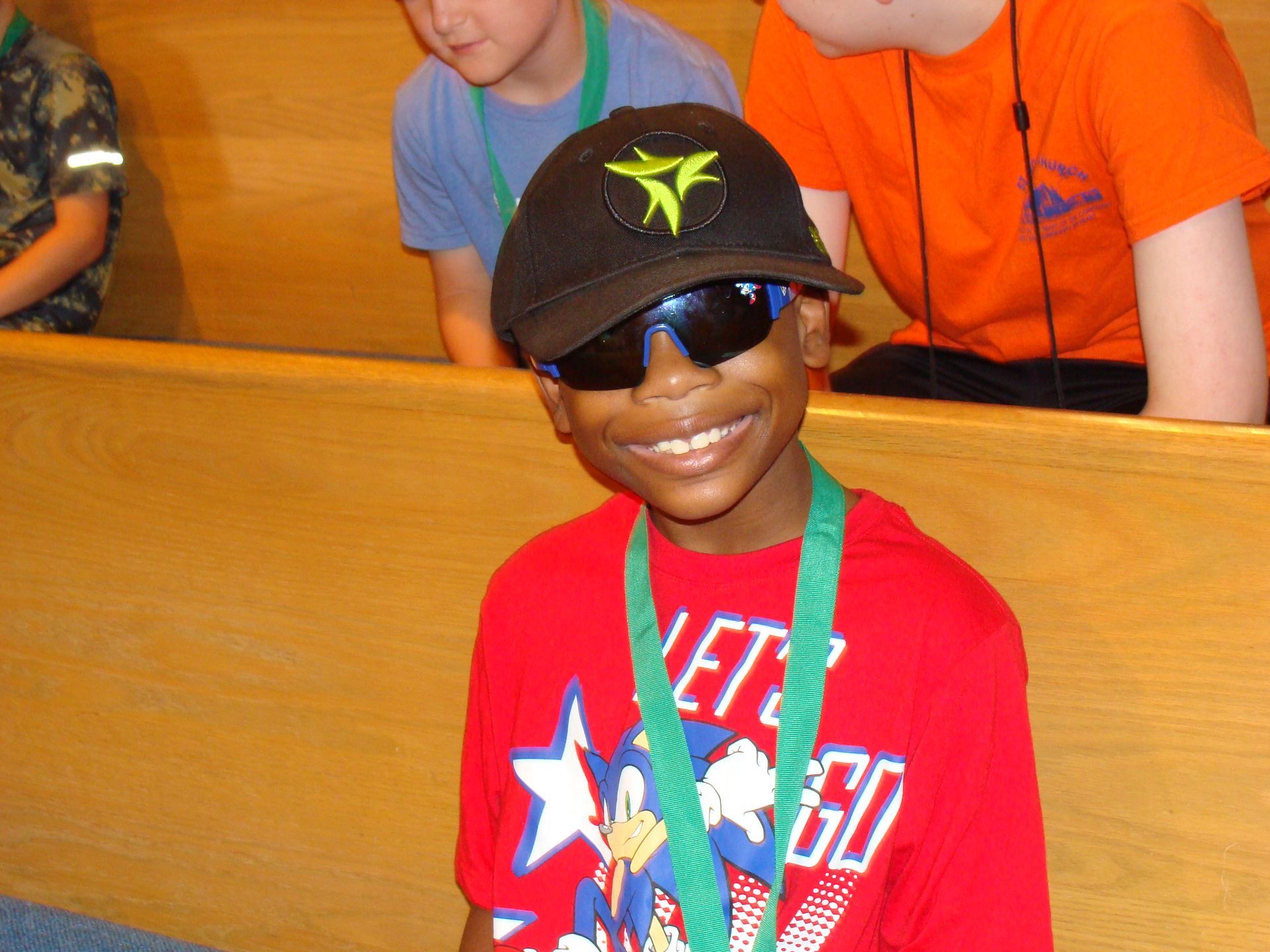 Smiling child in black cap and sunglasses, wearing a red shirt and green lanyard in a wooden bench setting