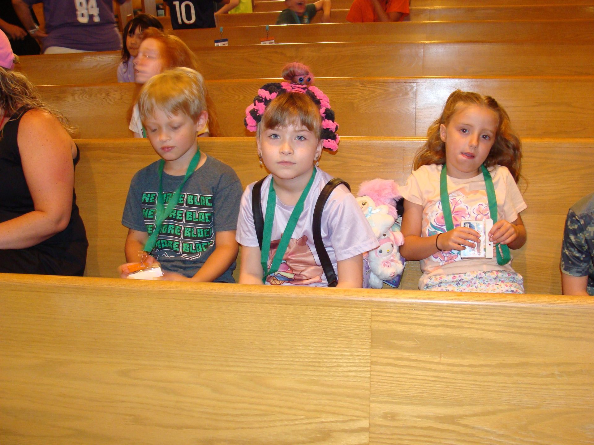 Three children sit on wooden bleachers; the middle child has pink hair bows and a stuffed toy behind her.
