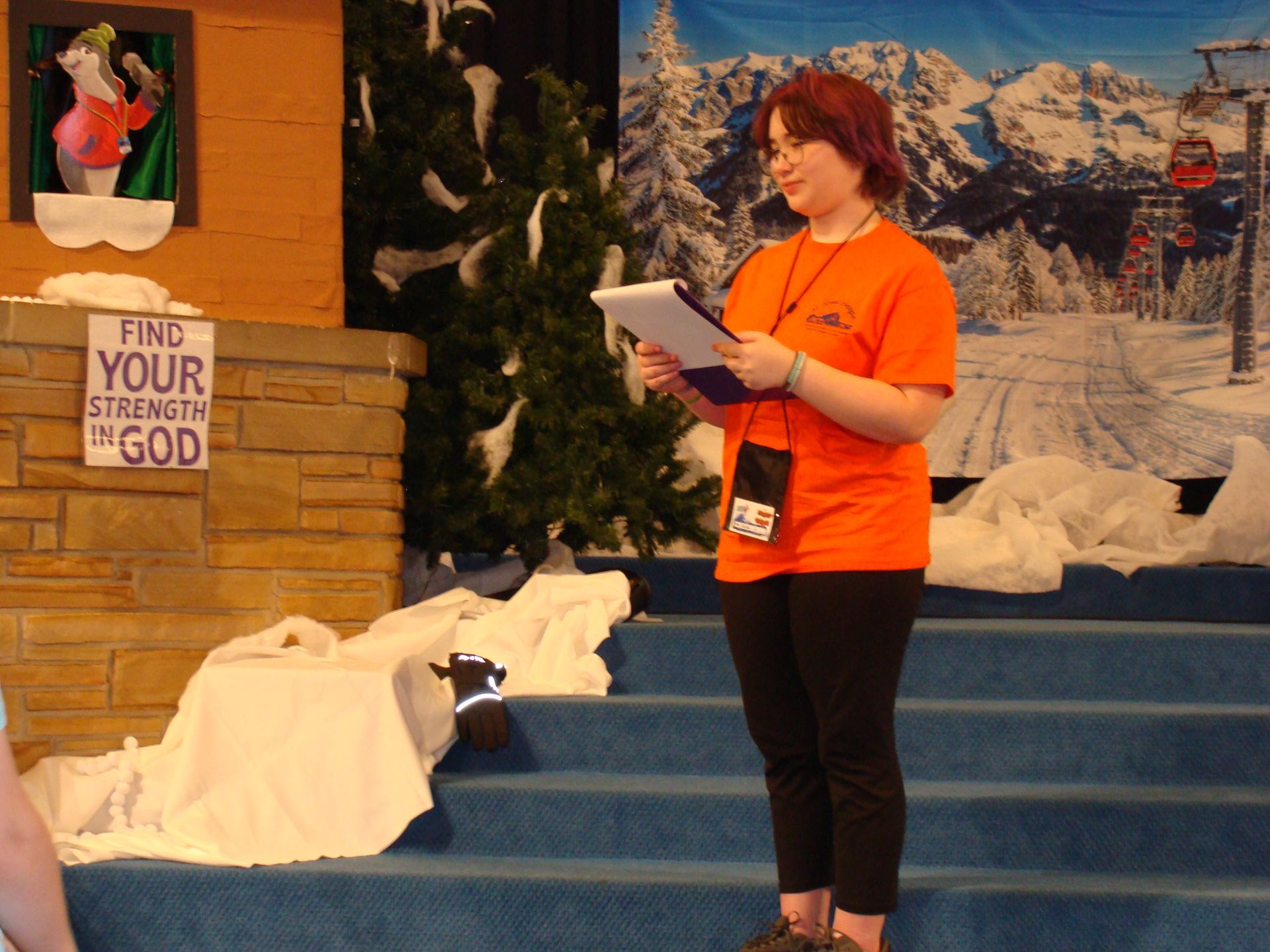 Woman in orange shirt reading on a snowy stage with winter backdrop and “Find your strength” sign
