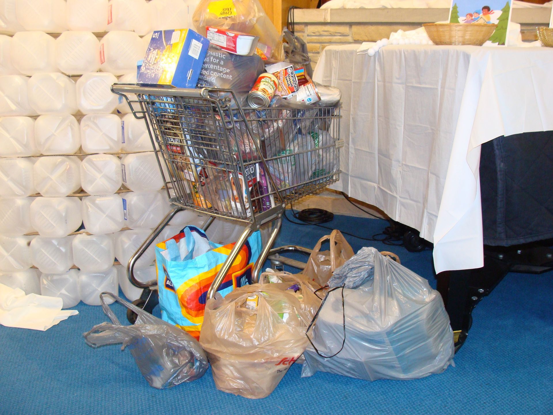 Shopping cart overflowing with groceries beside a stack of toilet paper rolls and plastic bags in a store room
