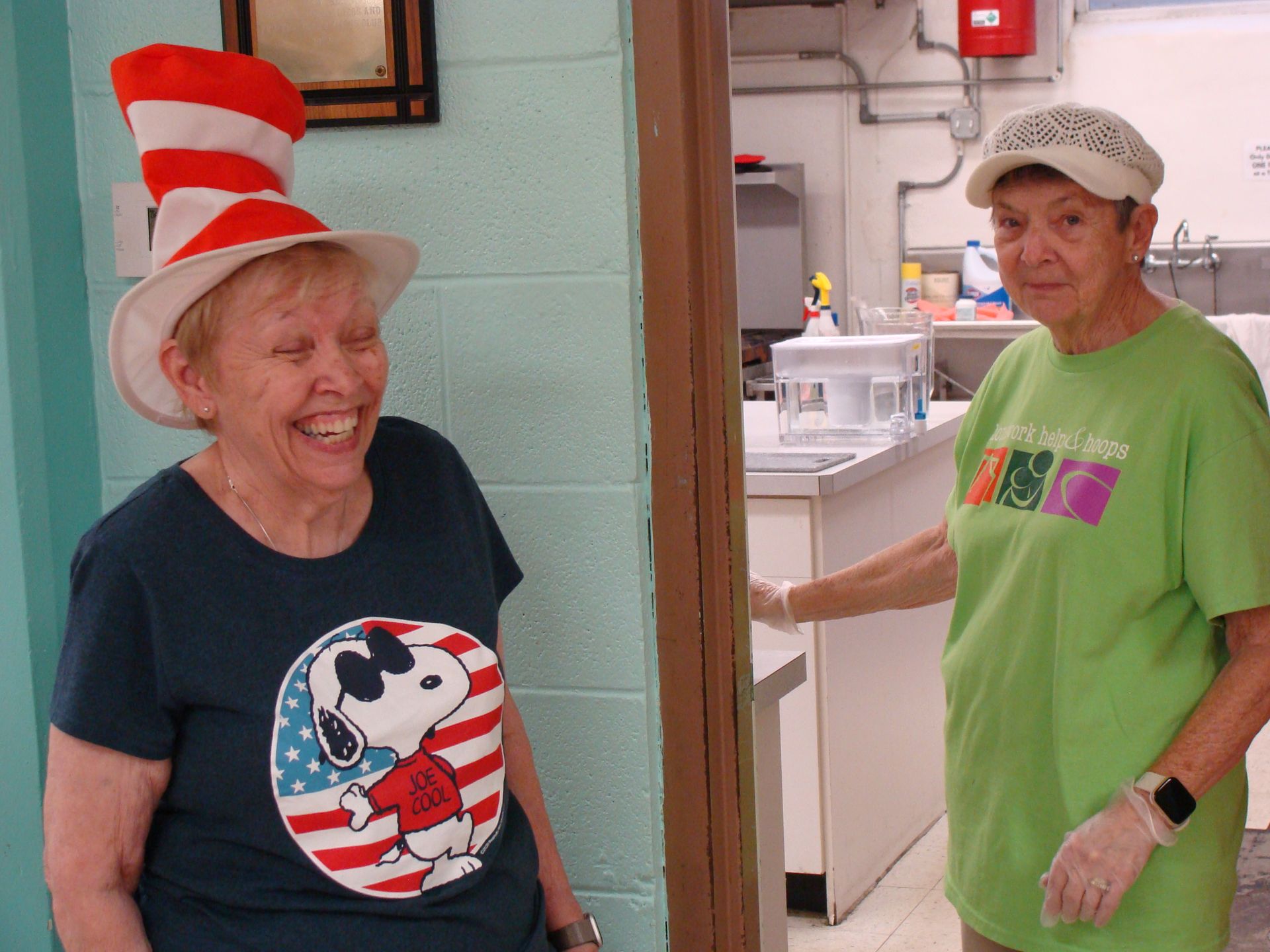 Two women smiling in a doorway, one in a red-striped hat and Snoopy shirt, the other in a green shirt