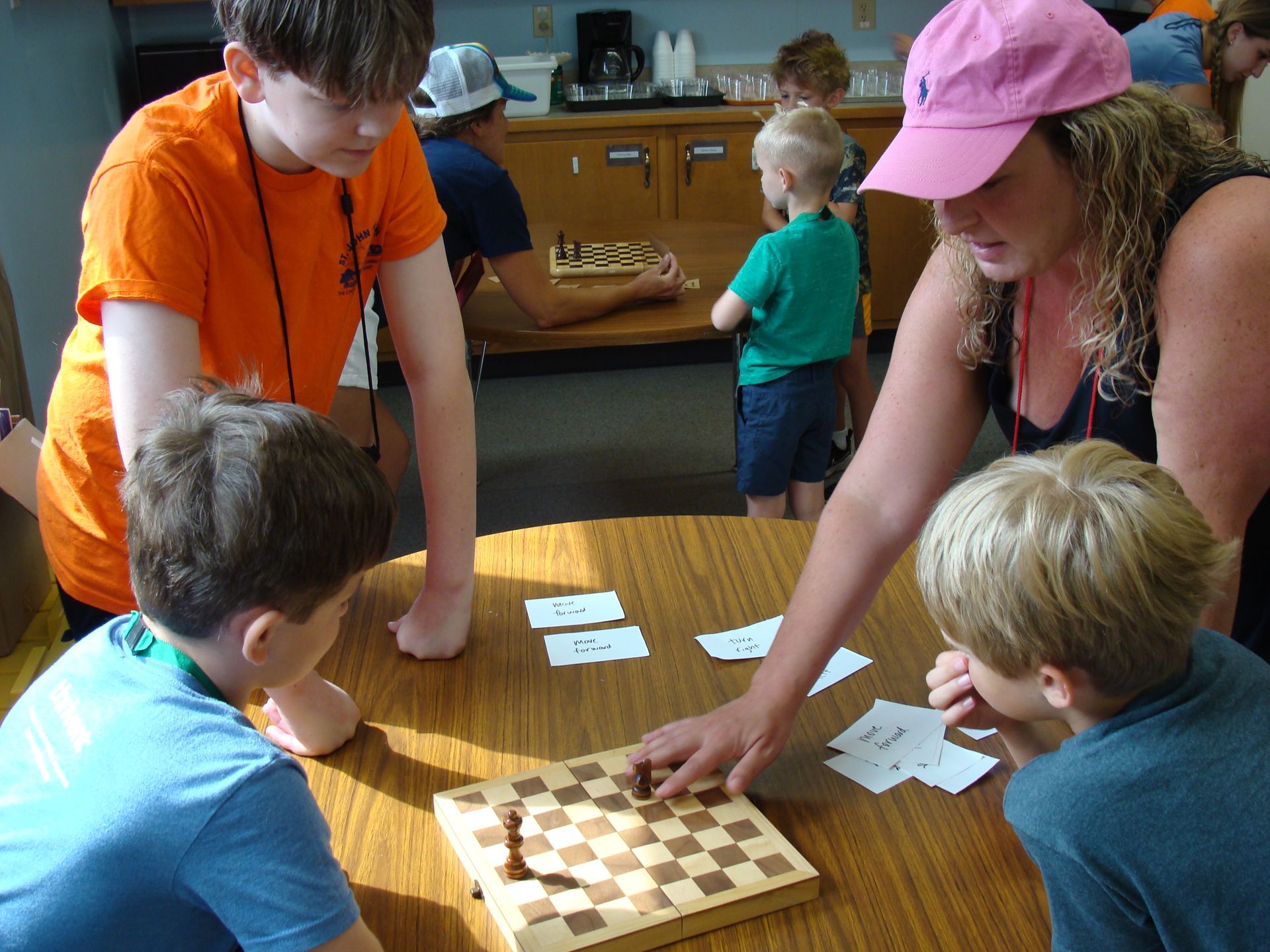People gathered around a chessboard on a table, with two adults and children playing in a room.