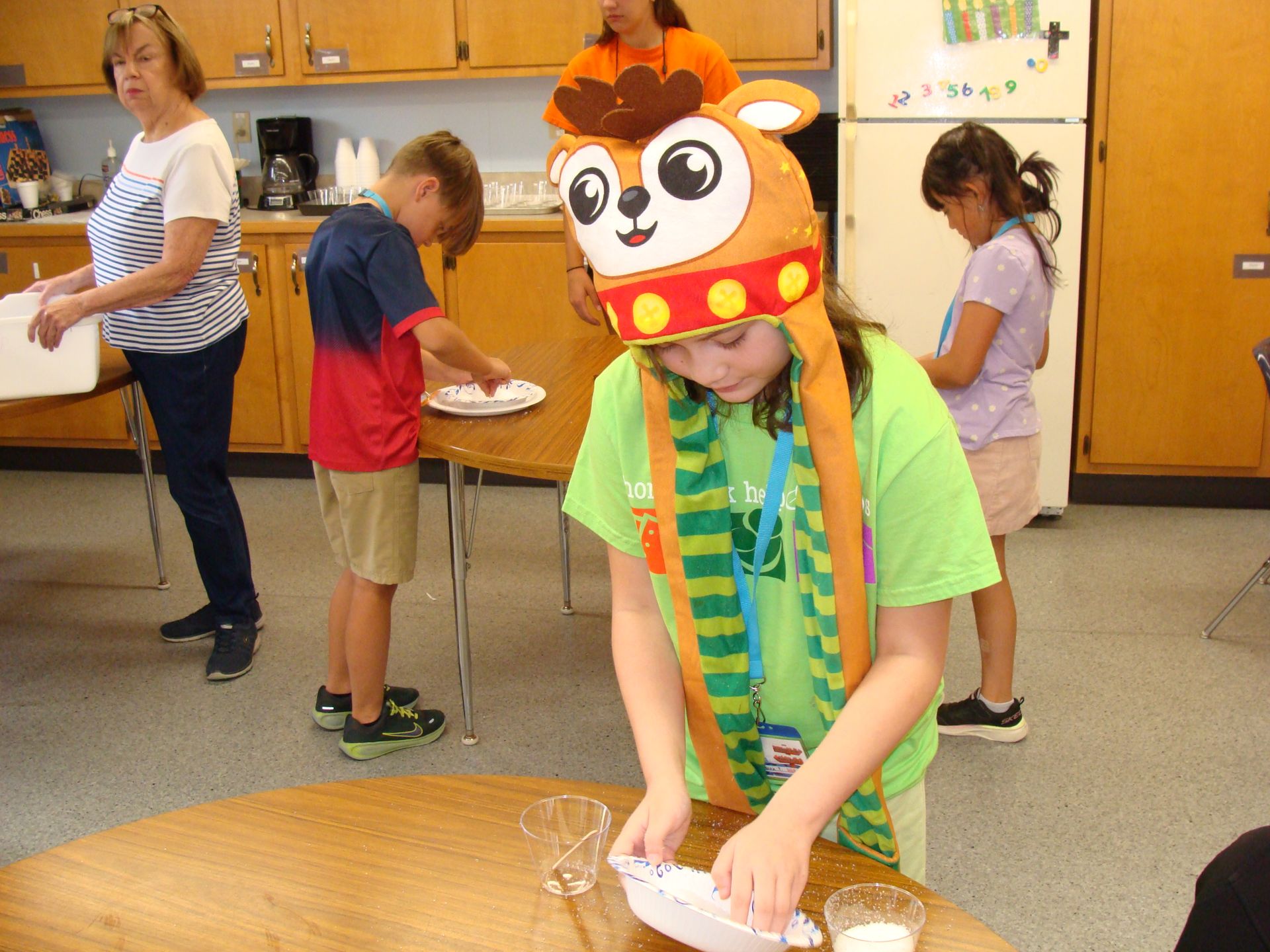 Children doing a classroom activity; one child wears a panda hat and writes at a table while others stand nearby.