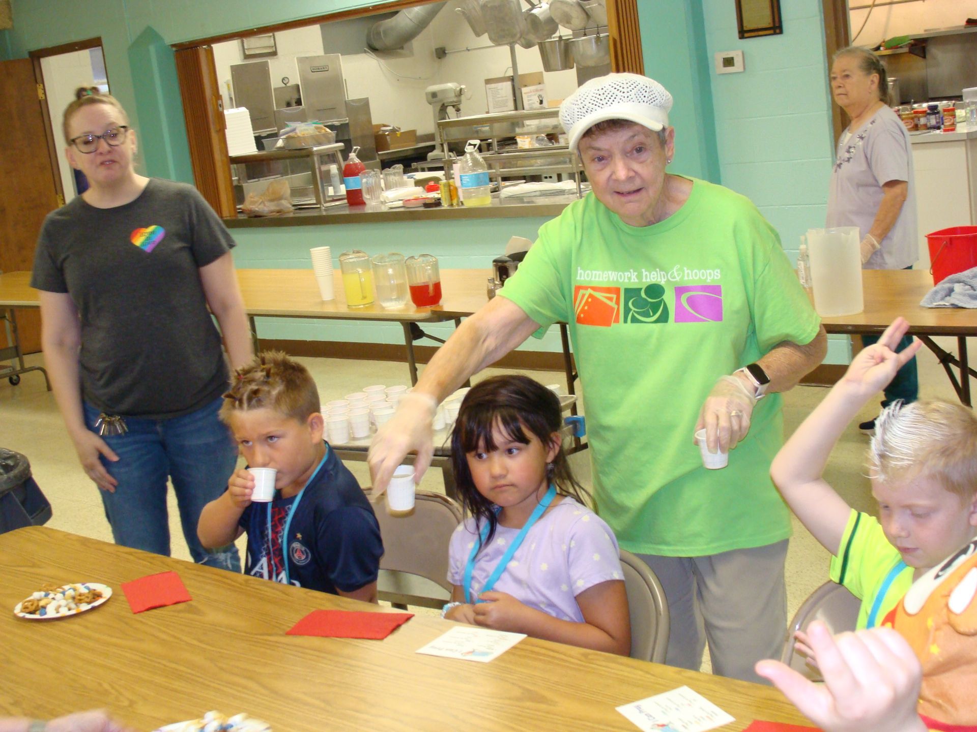 Adult guiding children in a classroom art activity at a table, with colorful supplies and paintings in the background
