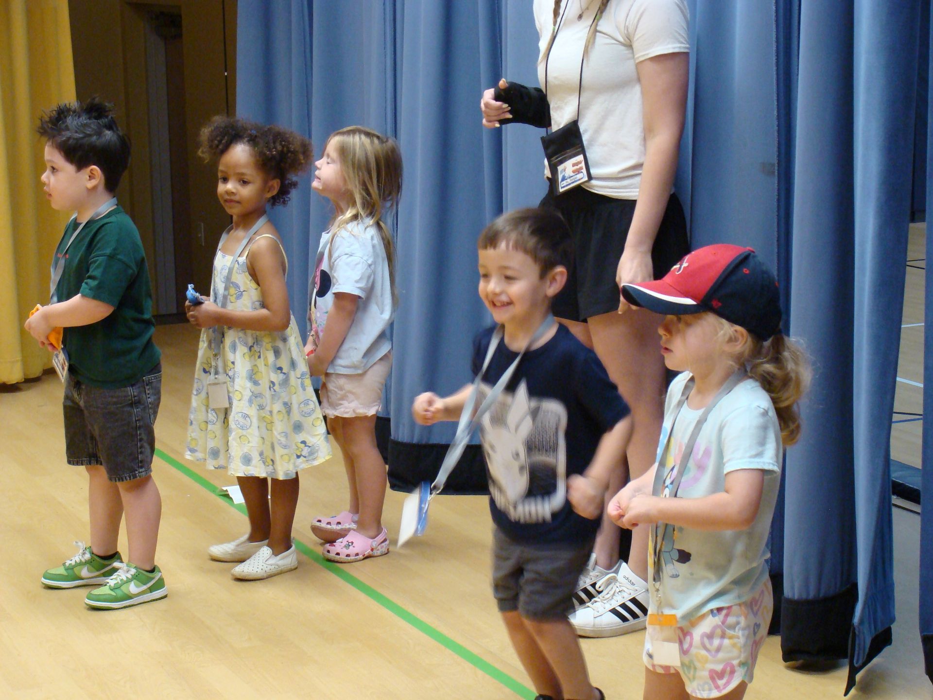 Children standing in a line on a stage, with an adult nearby and blue curtains behind them