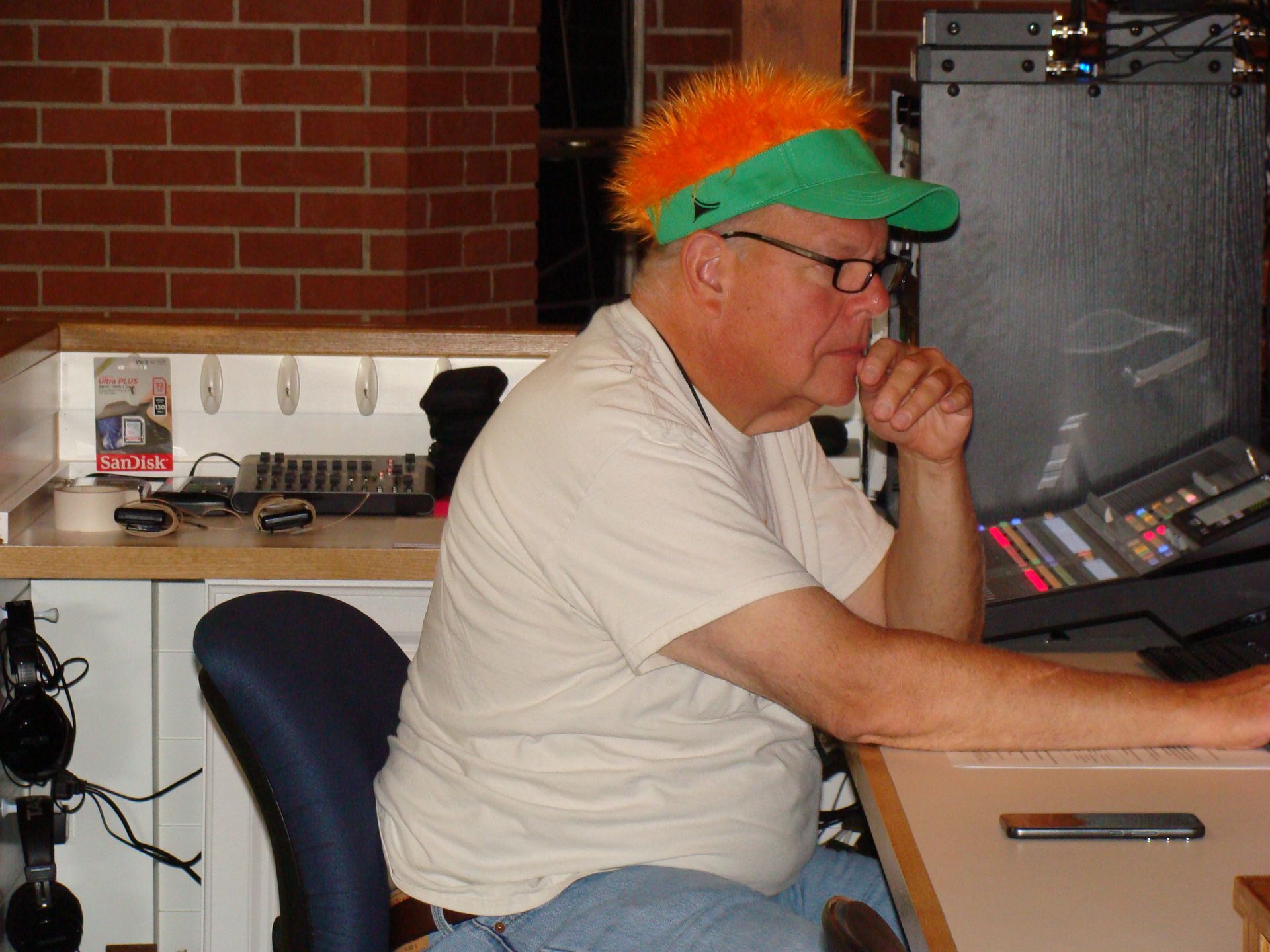 Person with orange hair and green visor sits at a control desk, resting chin on hand in a studio.