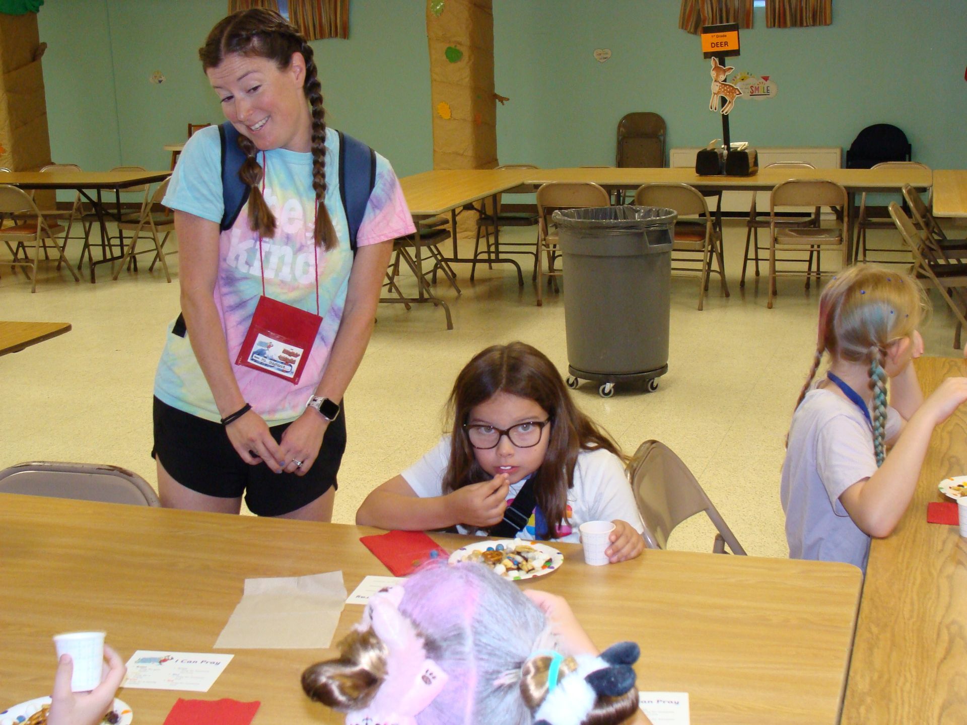 Woman stands beside seated children crafting at a table in a community room.