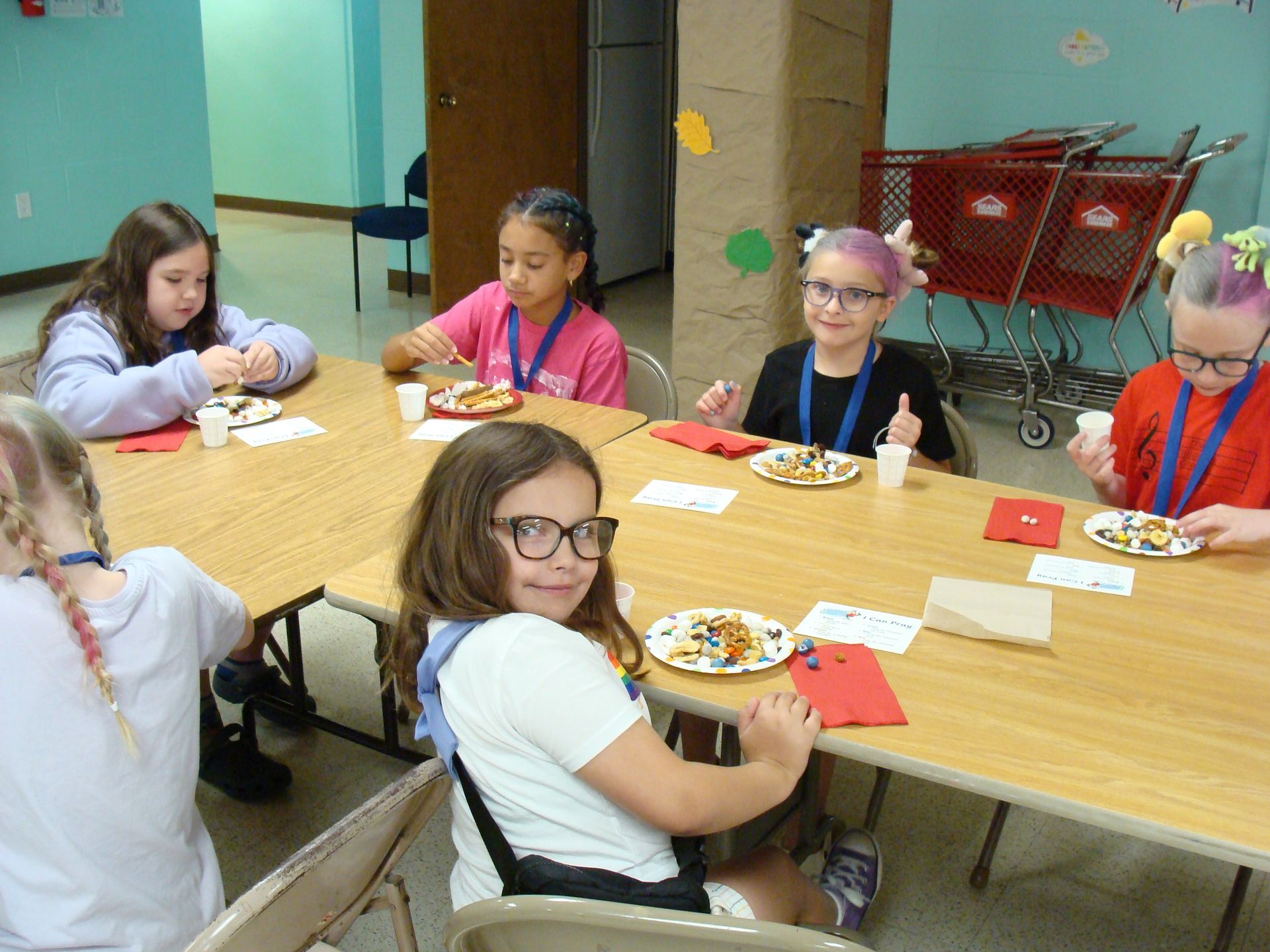 Children seated at a table in a classroom, eating and chatting during a snack or meal activity