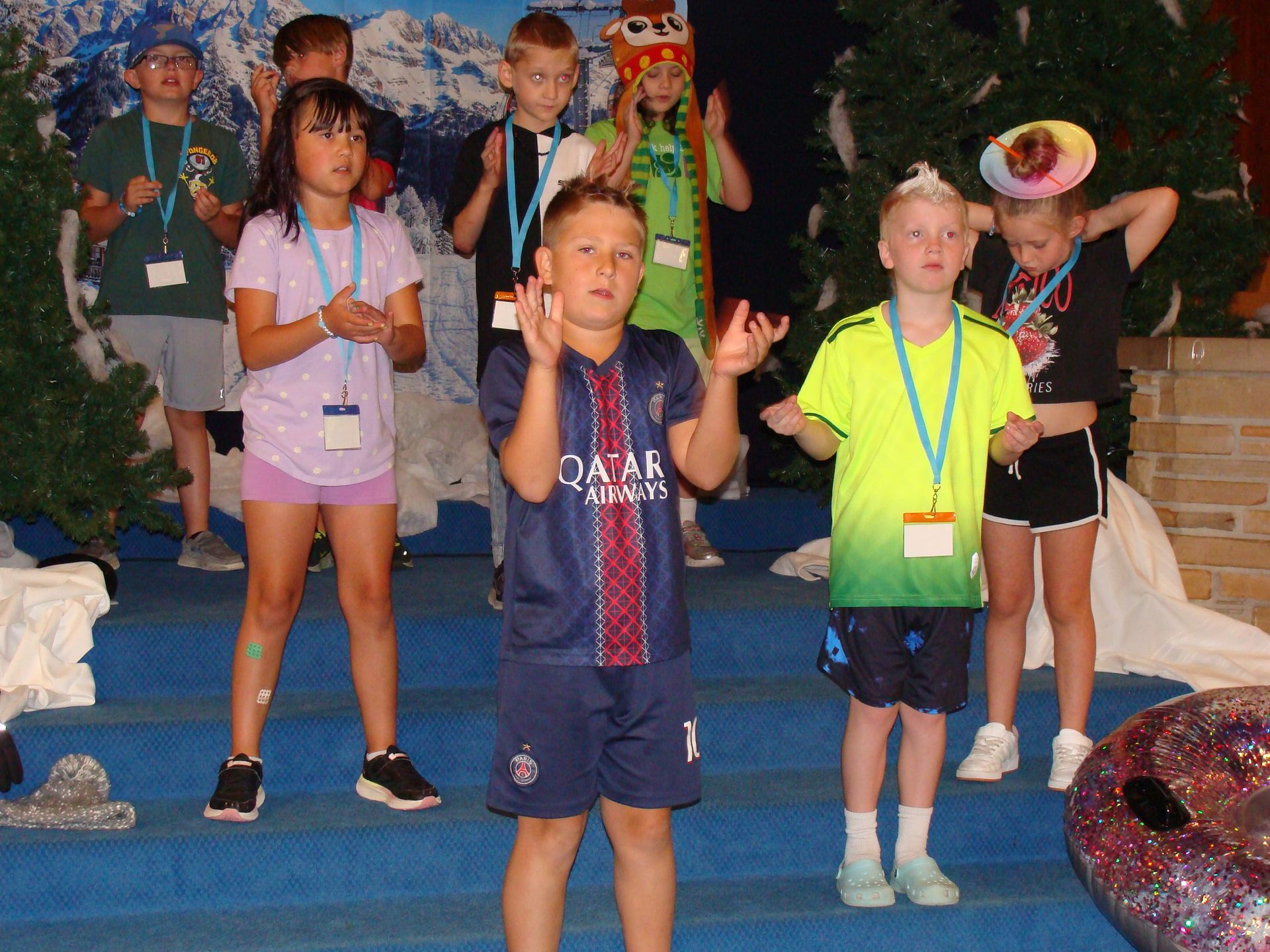 Children posing on a stage, wearing colorful costumes and lanyards, with one boy holding up a finger in front.