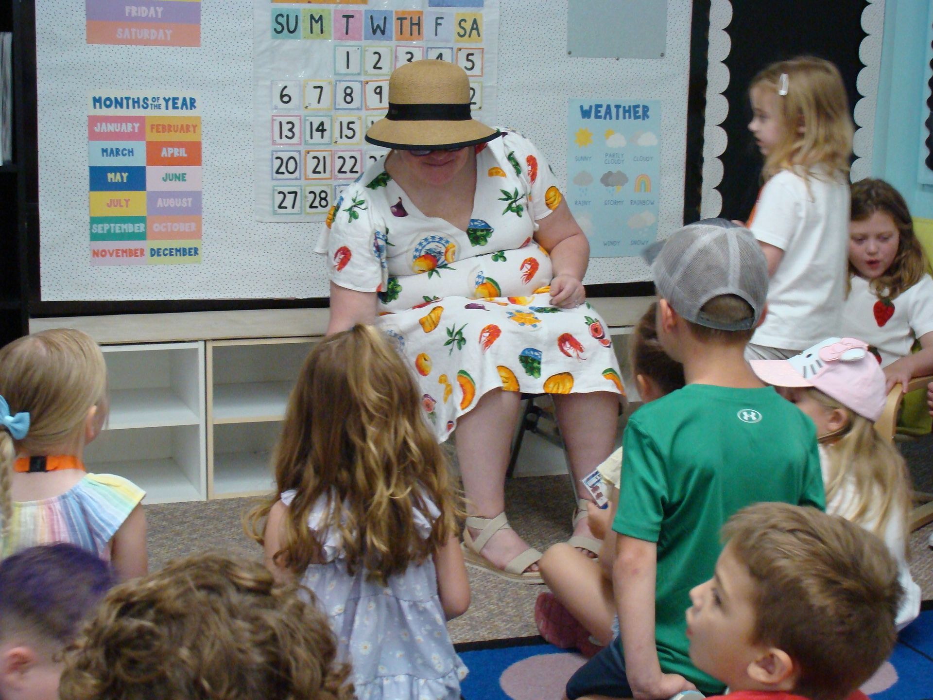 Teacher reading to children in a classroom, seated by a wall calendar and colorful alphabet chart.