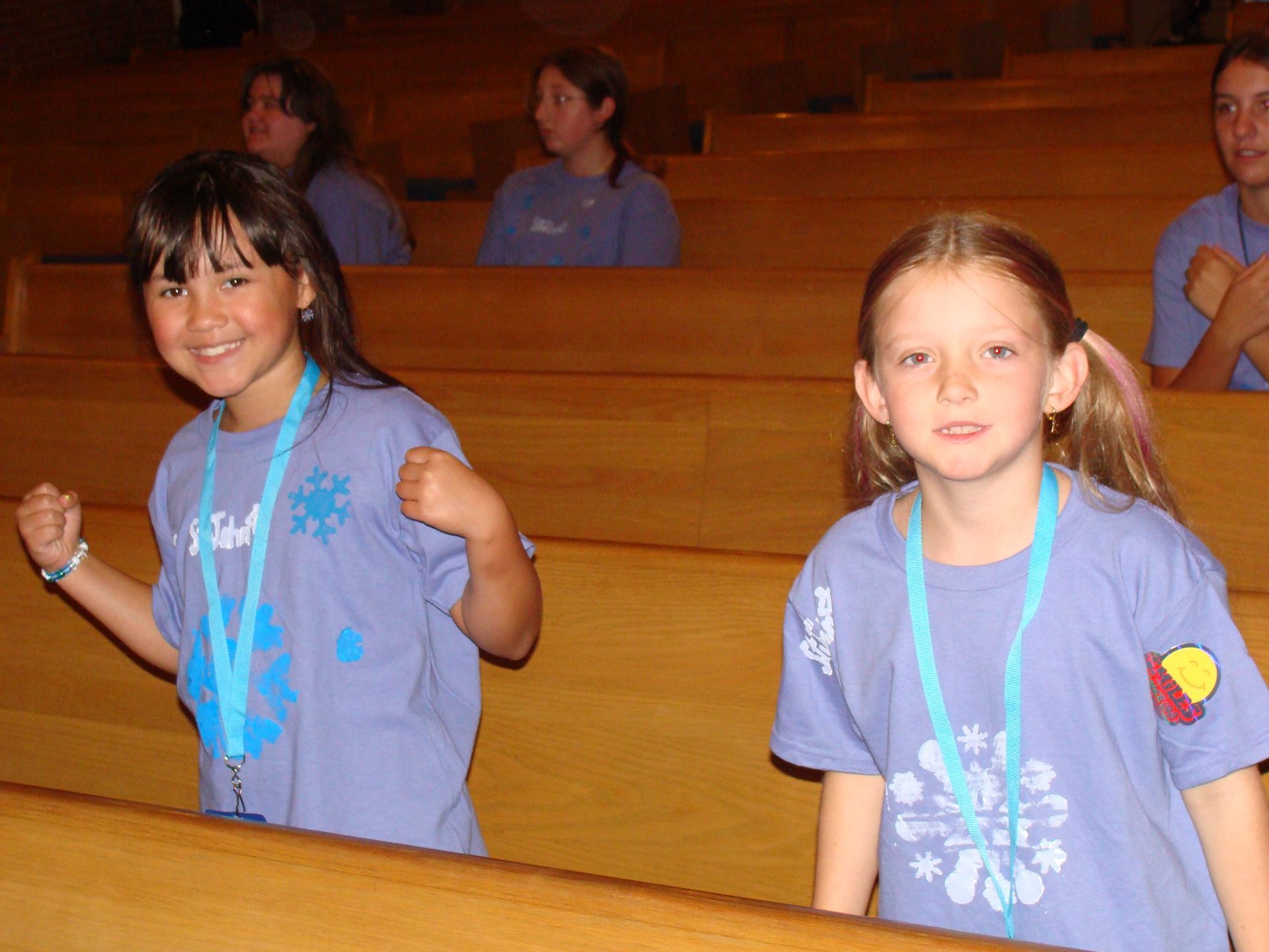 Two children smiling in matching purple shirts and blue lanyards, standing in a wooden auditorium.