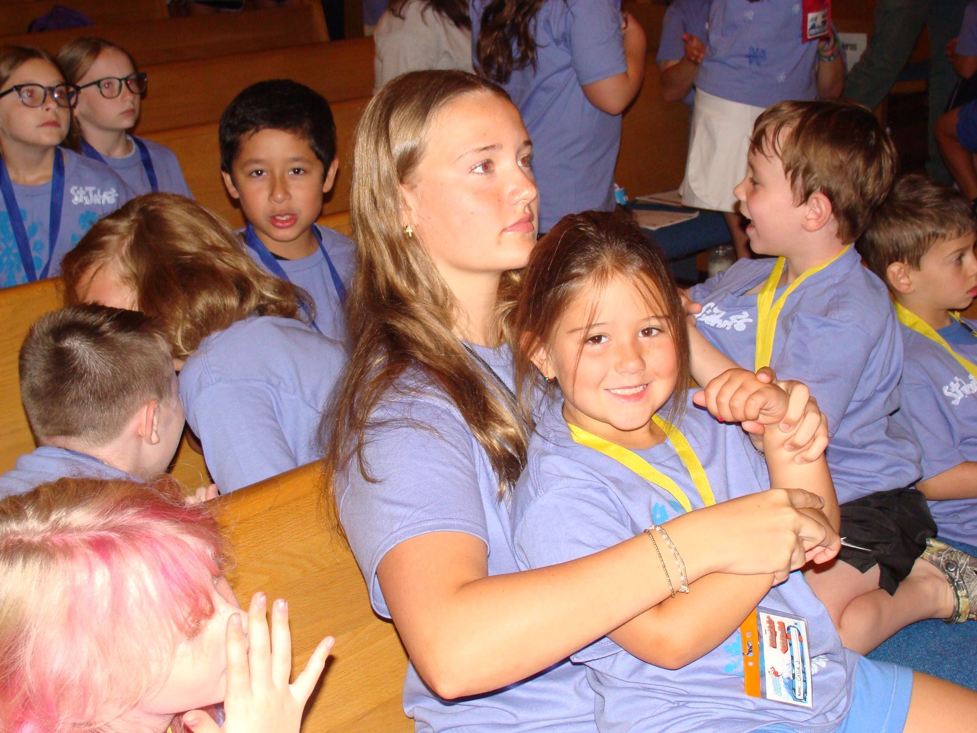 Children seated in a classroom, smiling and talking while wearing blue shirts and lanyards.