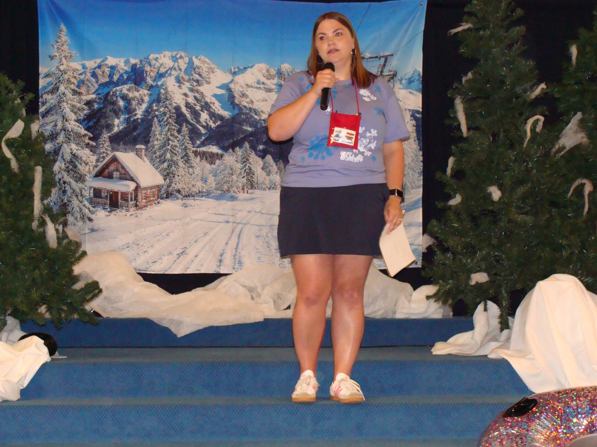 Woman speaking into a microphone on a stage with a snowy mountain backdrop and decorated evergreens