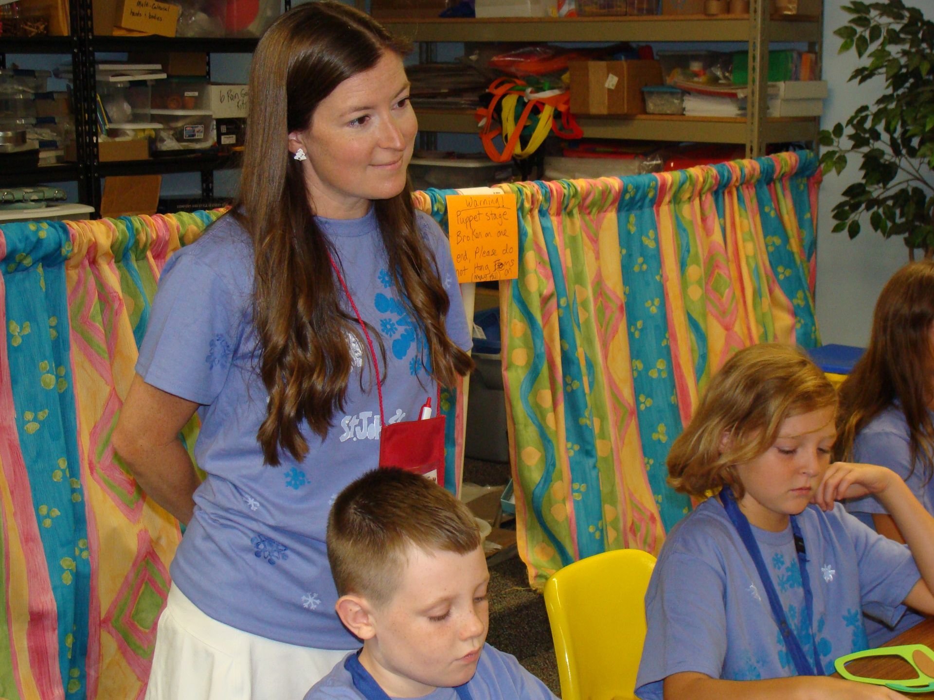 Woman stands behind seated children in a colorful classroom, with craft supplies and striped fabric in the background.