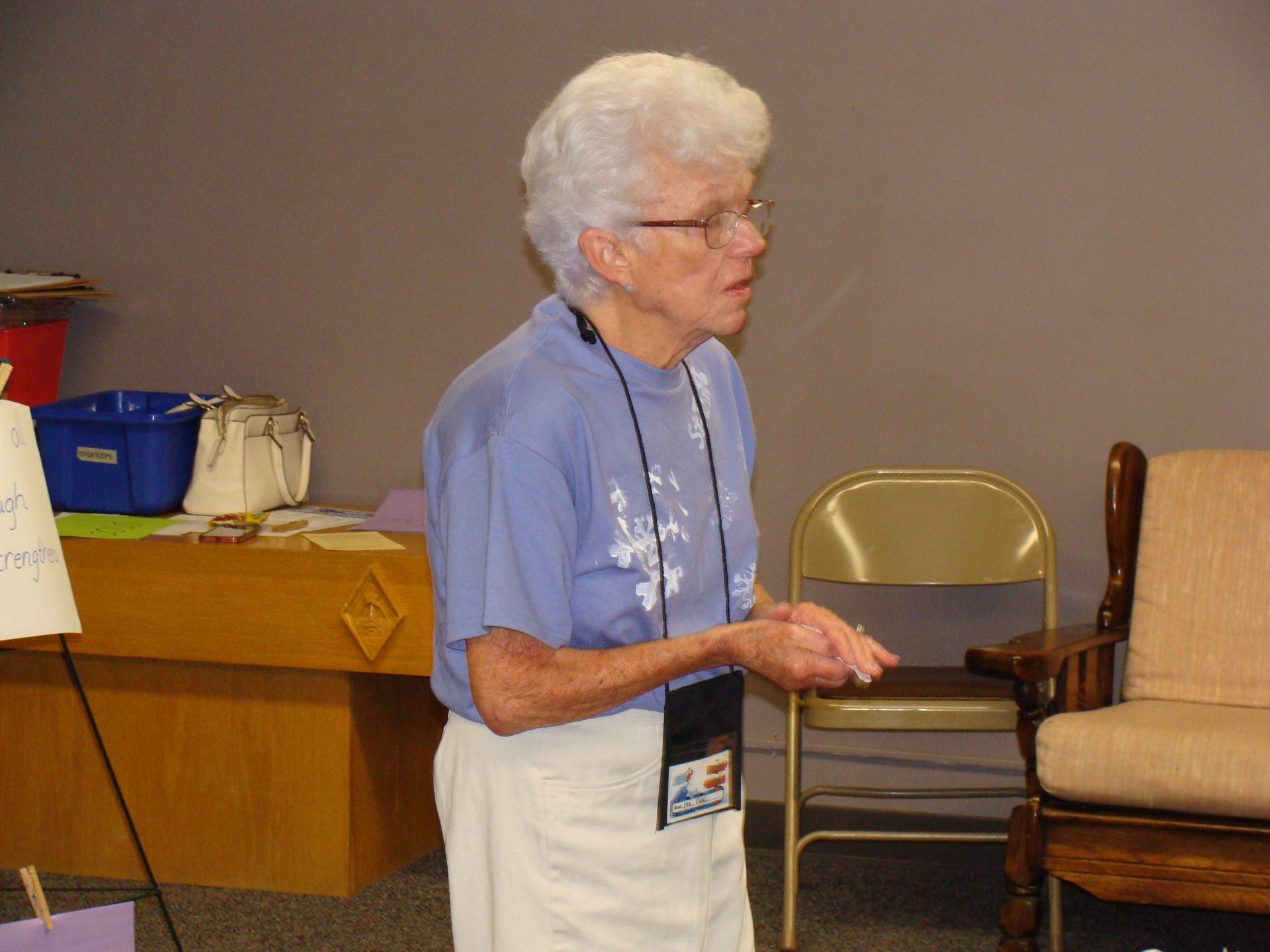 Woman in a blue shirt standing beside chairs in a room, holding papers and speaking.