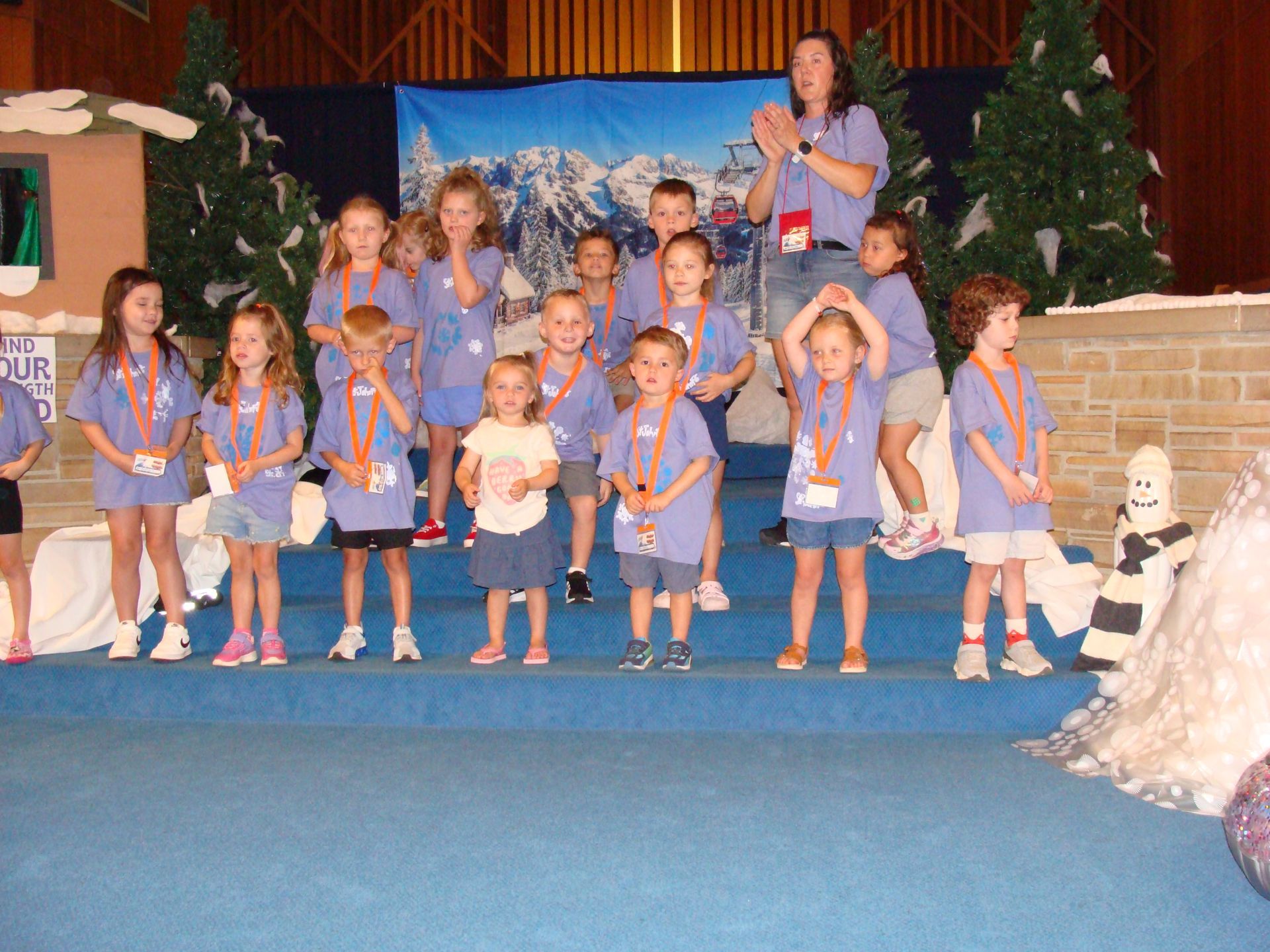 Children in blue shirts pose on a stage with a teacher, greenery, and a brick wall backdrop