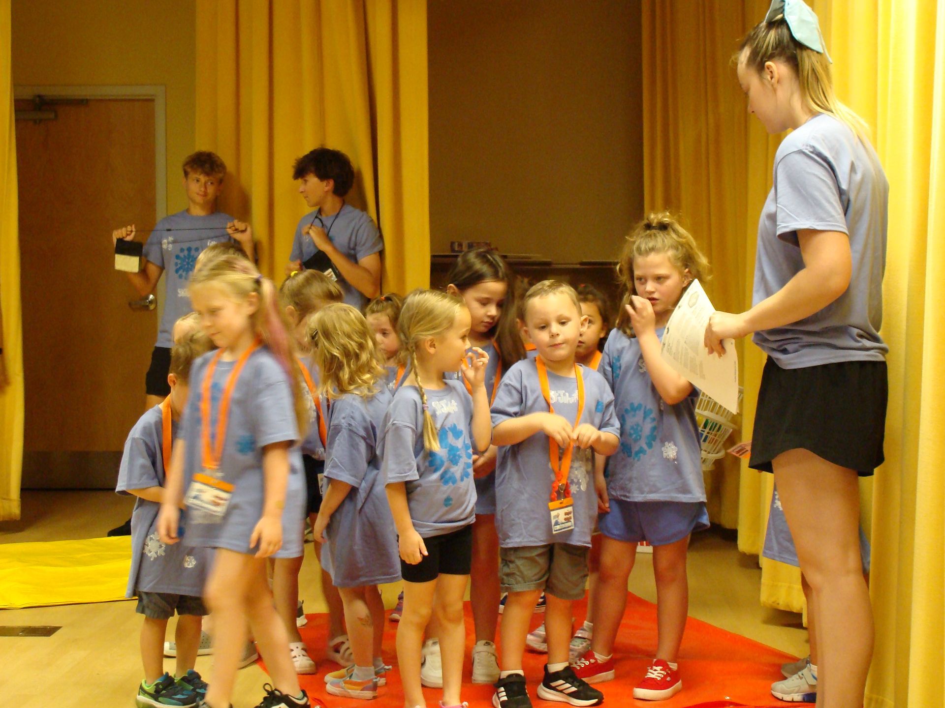 Children in blue shirts and orange shorts stand on a stage while an adult supervises nearby.