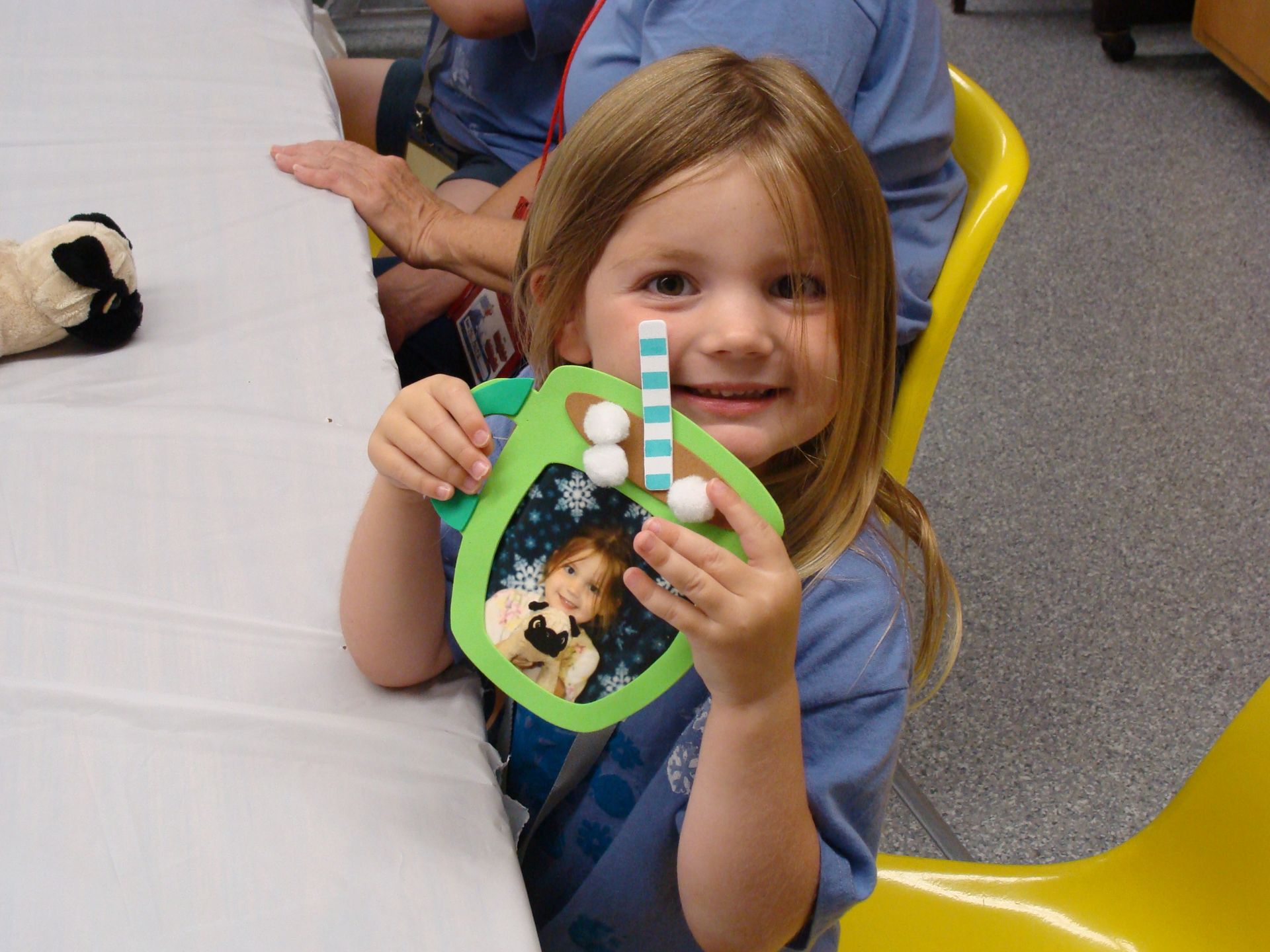 Child smiling and holding a green photo frame craft at a table in a classroom.