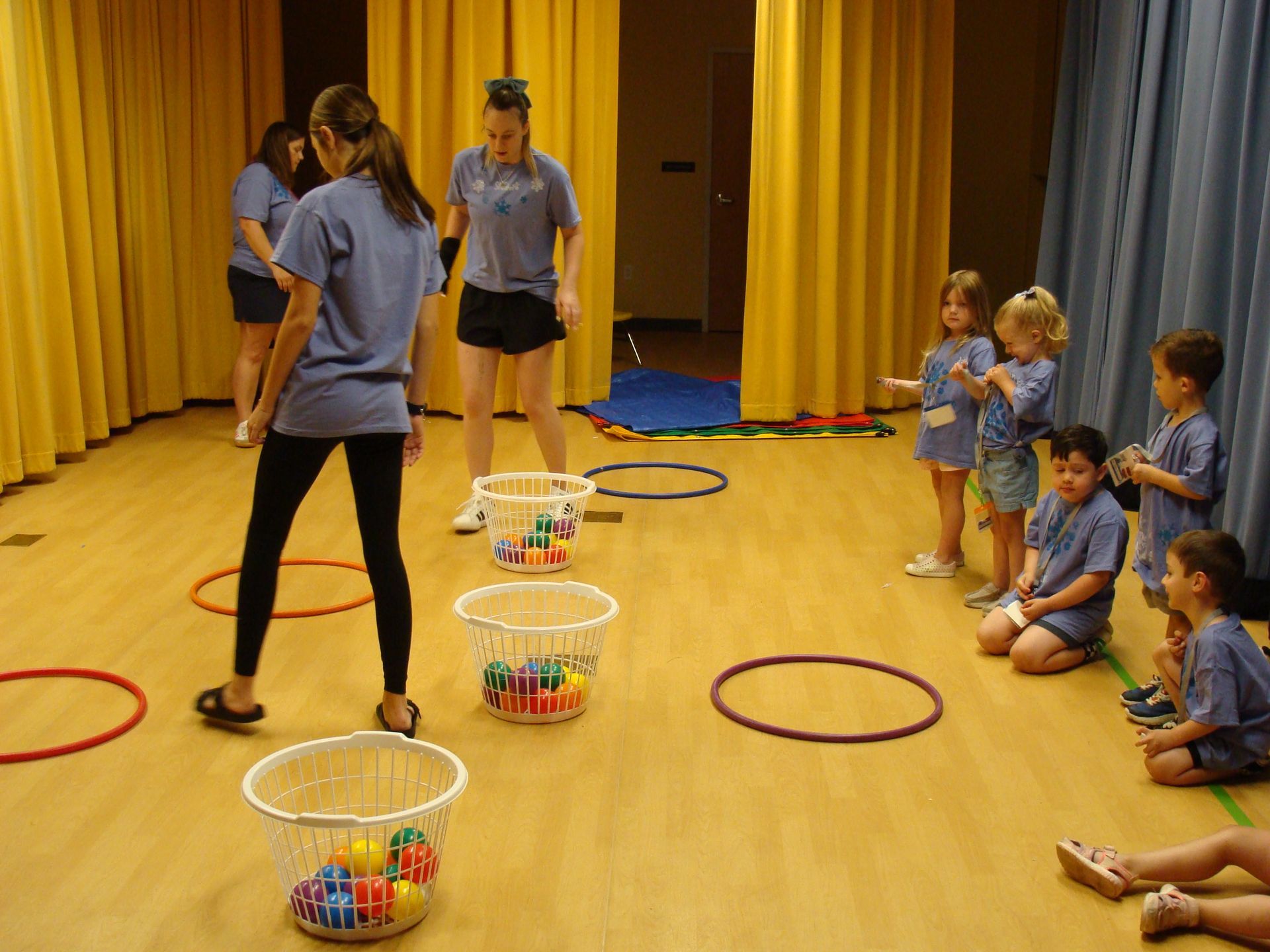 Children playing a ring toss game in a gym, with buckets and colorful rings on the floor