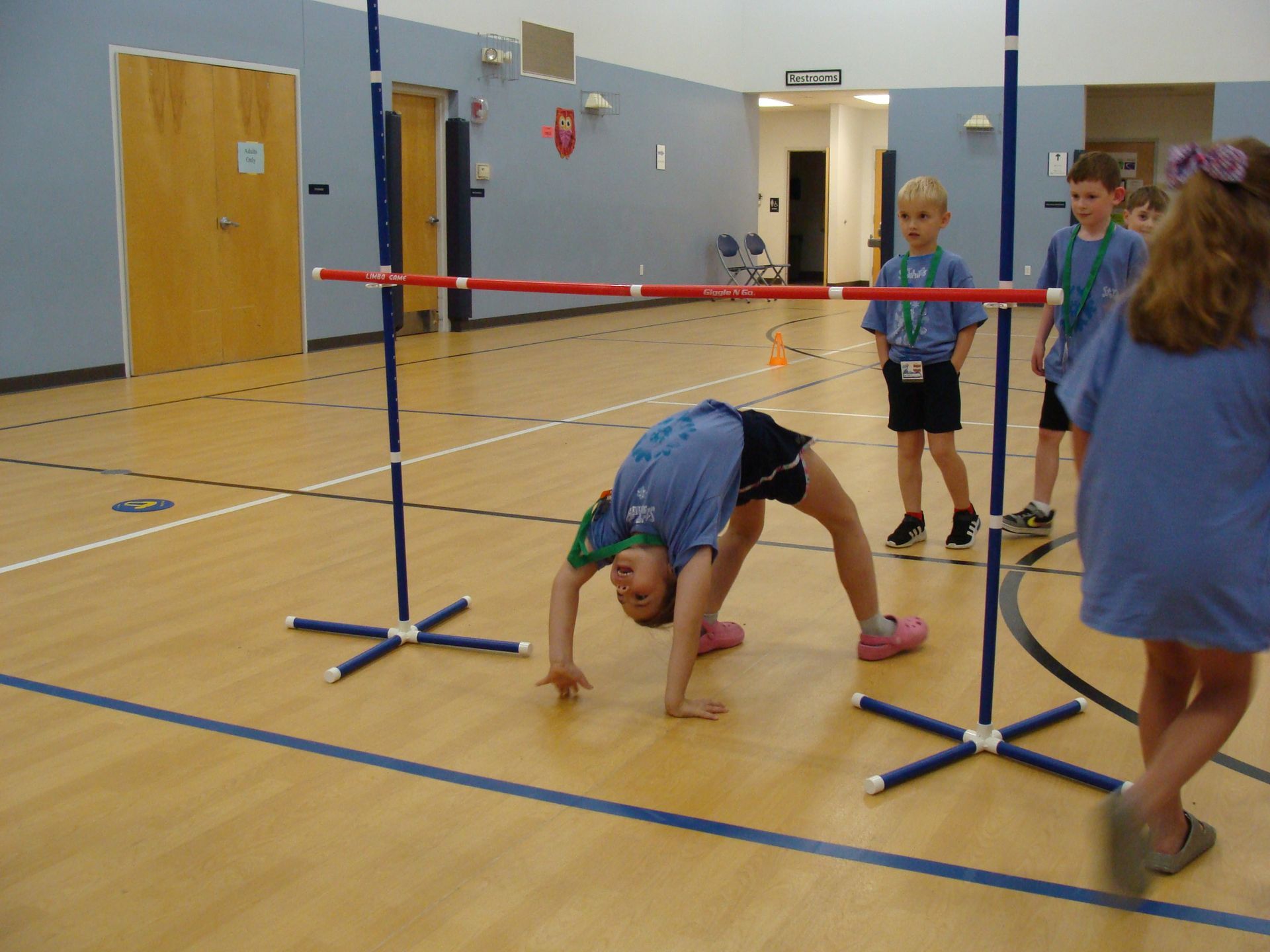 Children limbo under a red bar in a gymnasium, with one doing a backbend under it.