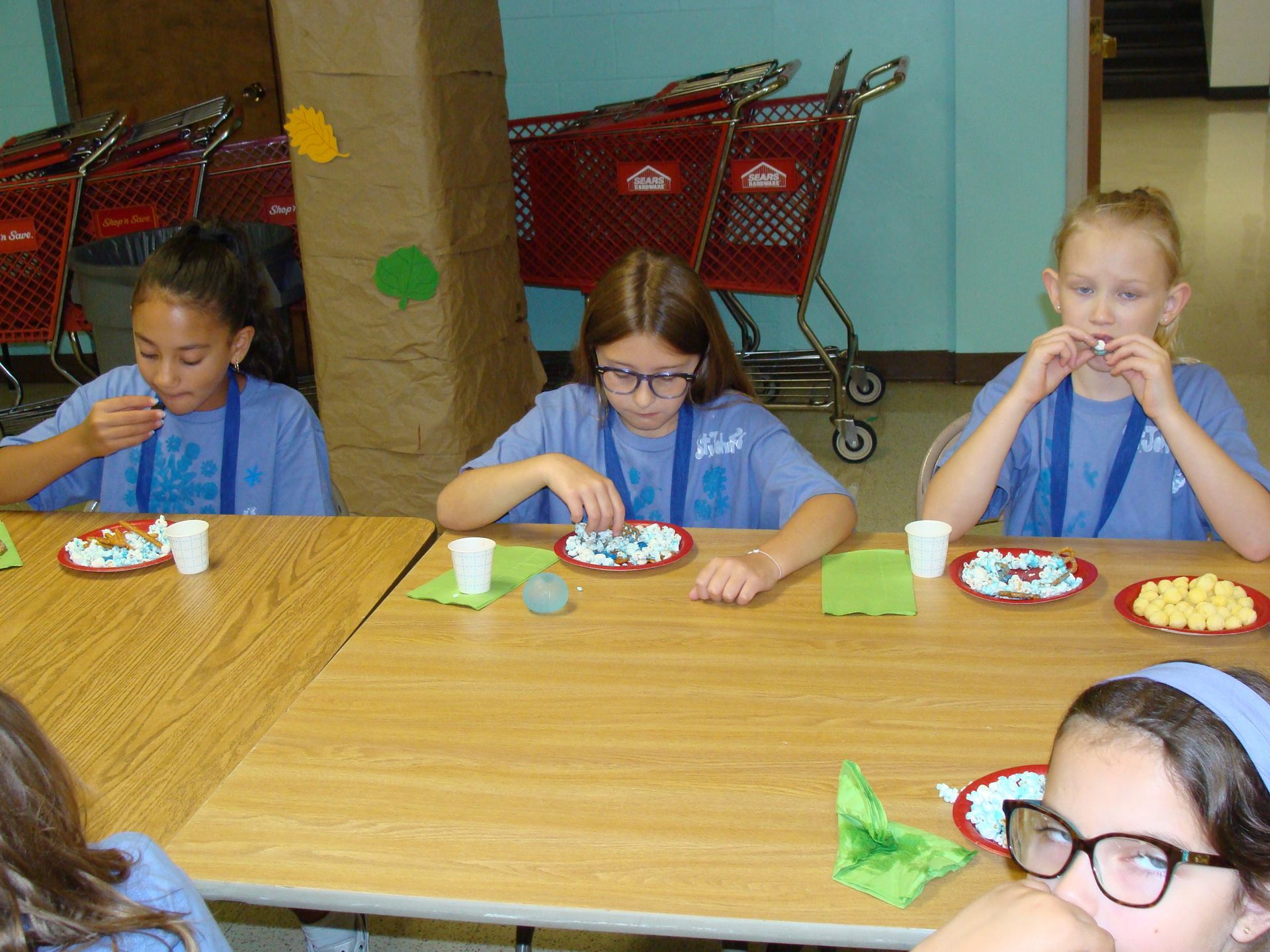 Children eating at a wooden table with paper plates and cups in a cafeteria or classroom setting