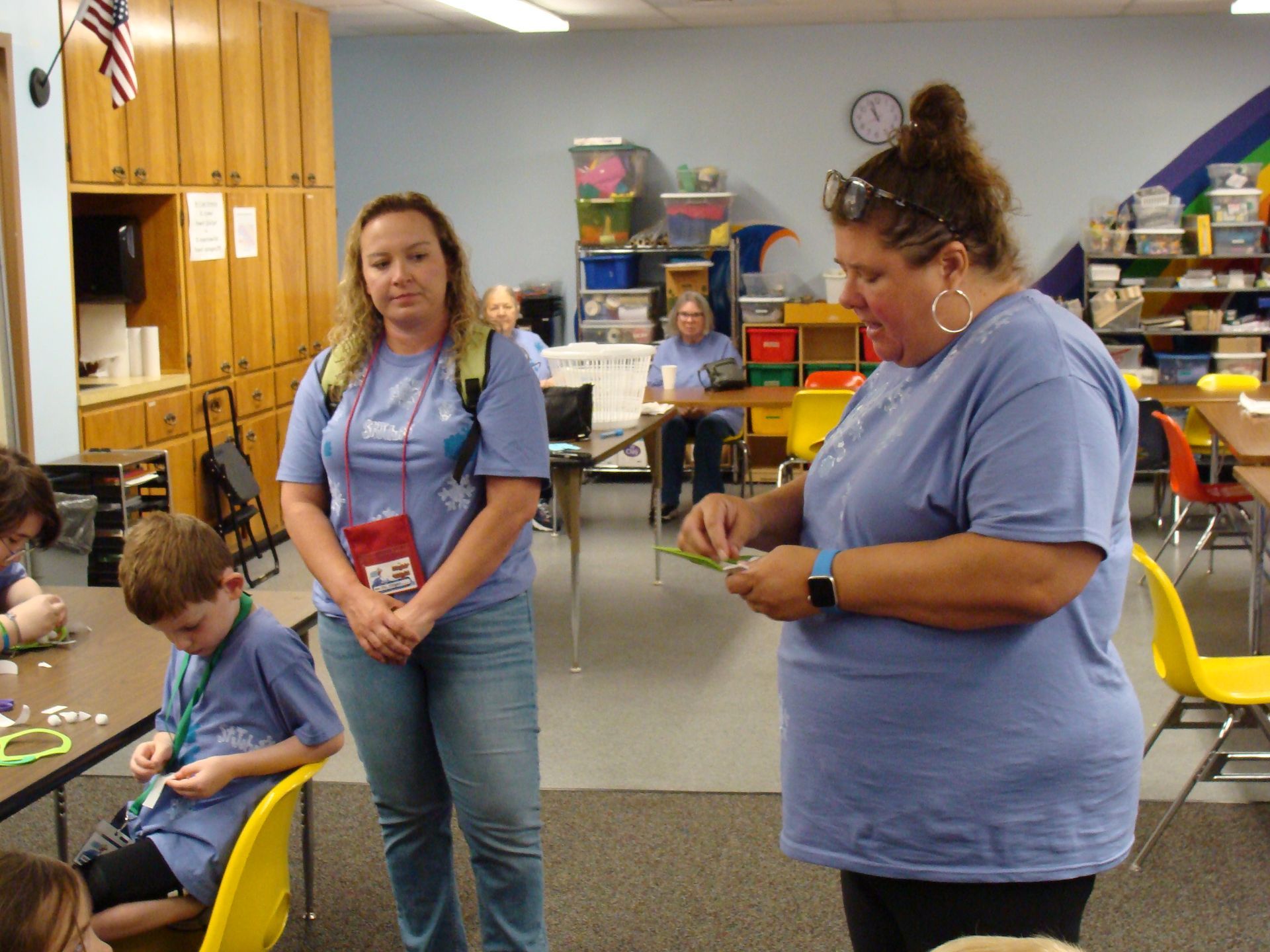 Two women standing in a classroom while children work at tables and on the floor.