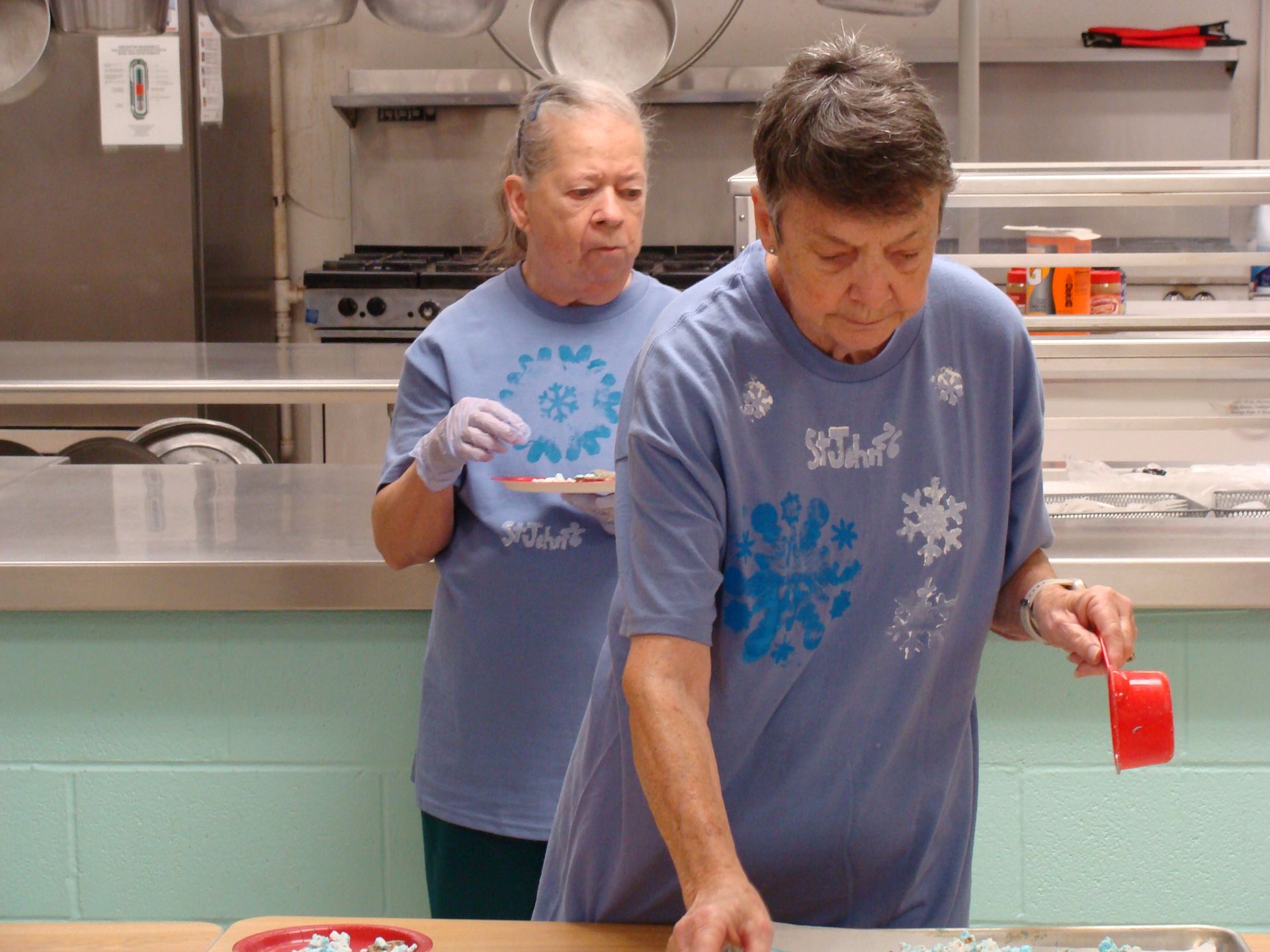 Two people in blue shirts stand at a stainless-steel counter, one holding a red cup in a kitchen-like setting.