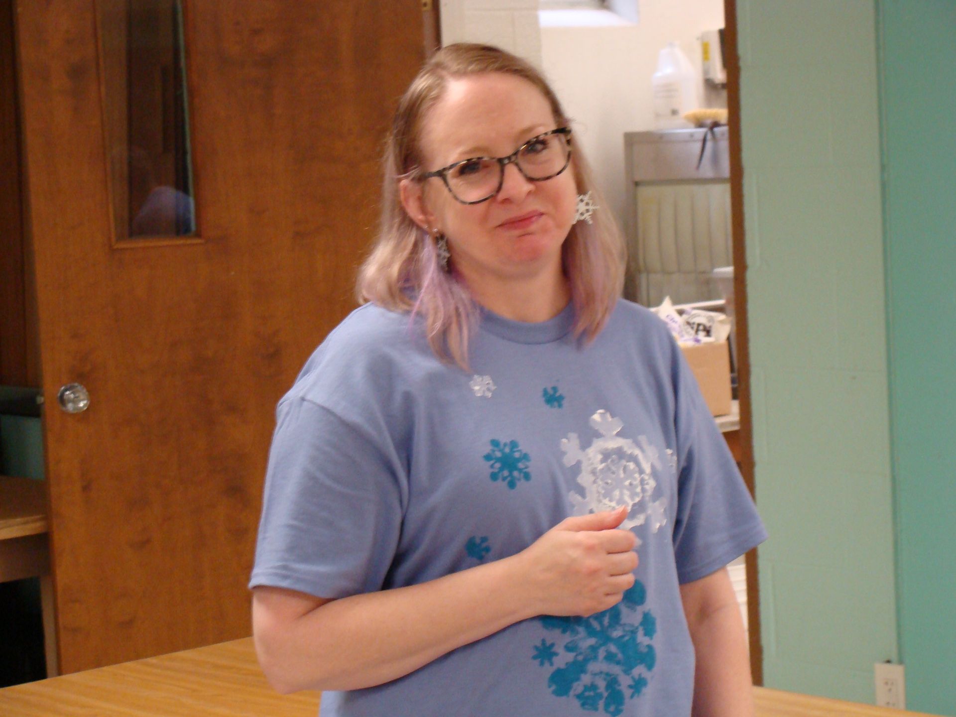 Person in a light blue shirt holding a drink, standing indoors near a wooden door.