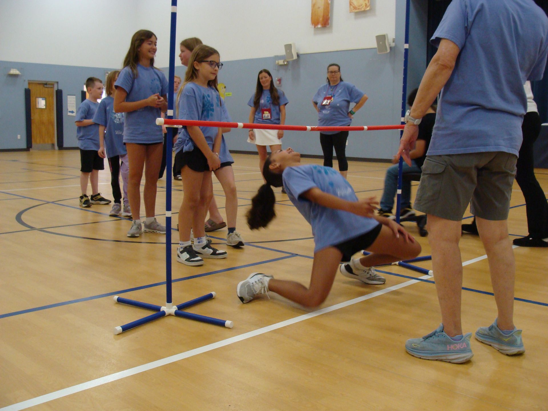 Students limbo under a red bar in a gymnasium during a physical education activity