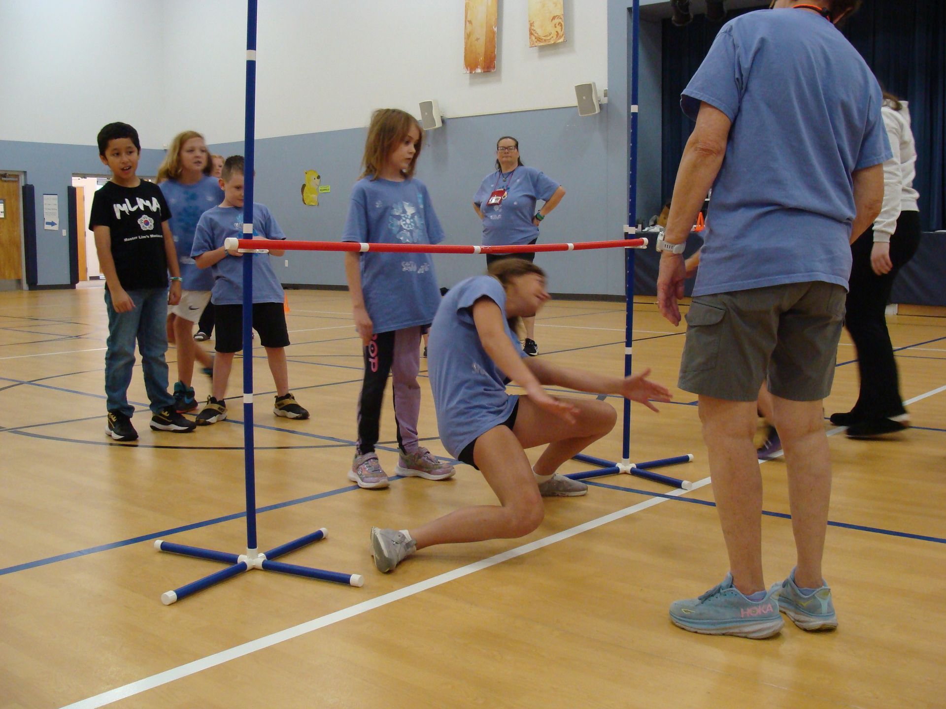 Children in a gym playing limbo under a red bar while one kneels to adjust it