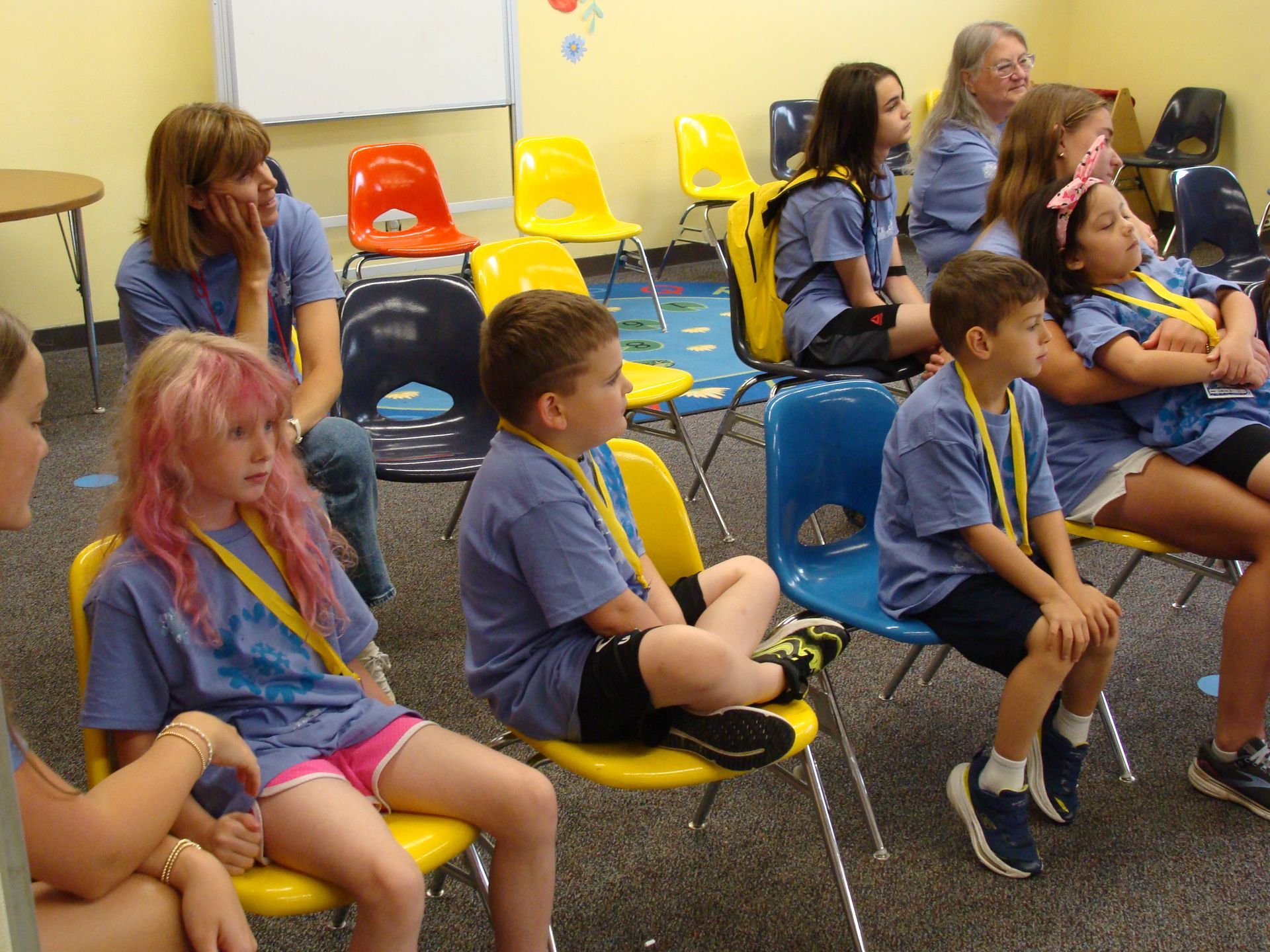 Children seated on colorful chairs in a classroom, facing forward during an activity.