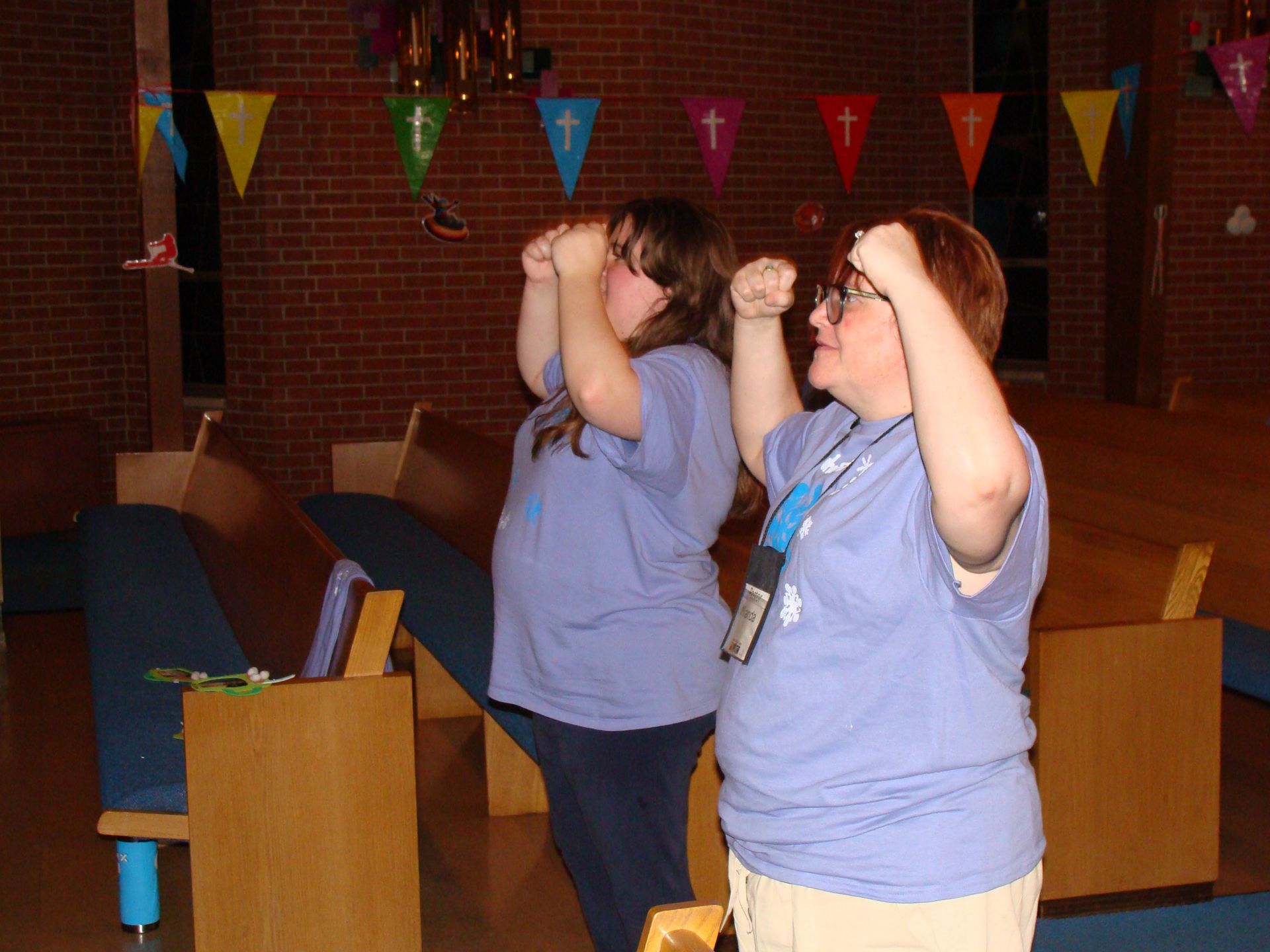 Two people in blue shirts raise their hands in a church or community hall with colorful banners overhead