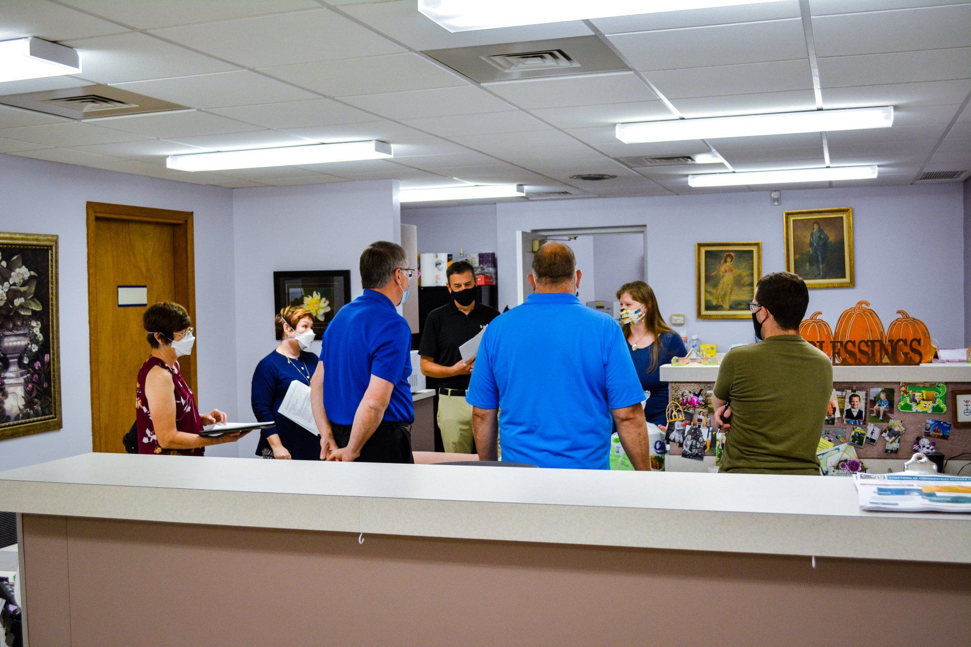 People standing at a counter in a bright office or gallery lobby, talking with a masked staff member.