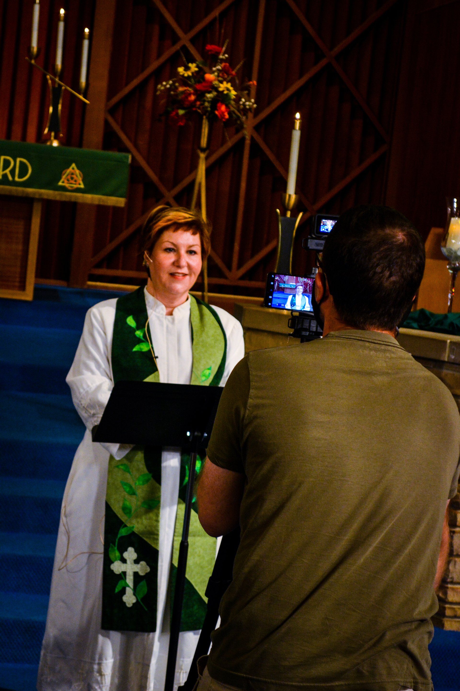 Woman in white robe with green stole speaking at a lectern while a man films her in a church.
