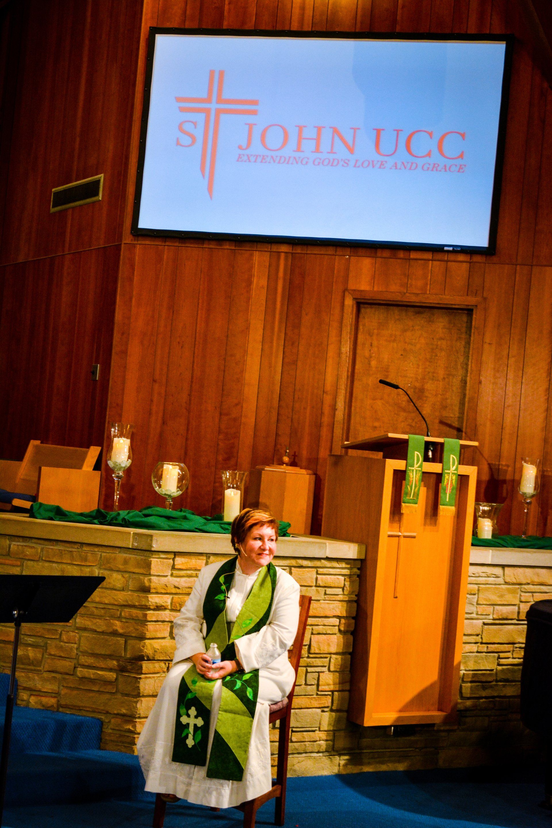 Person seated in a church sanctuary before a pulpit and “Join UCC” screen, wearing a white robe and green stole