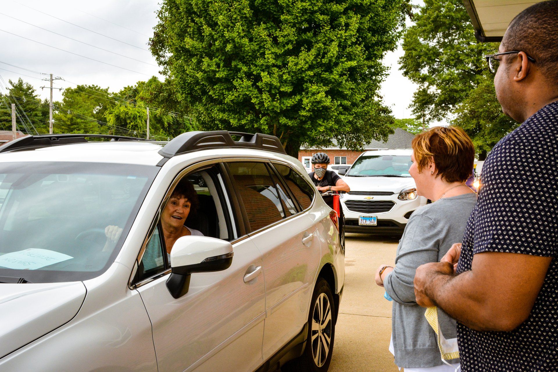 Car at curbside with driver speaking to two people near a tree-lined street.