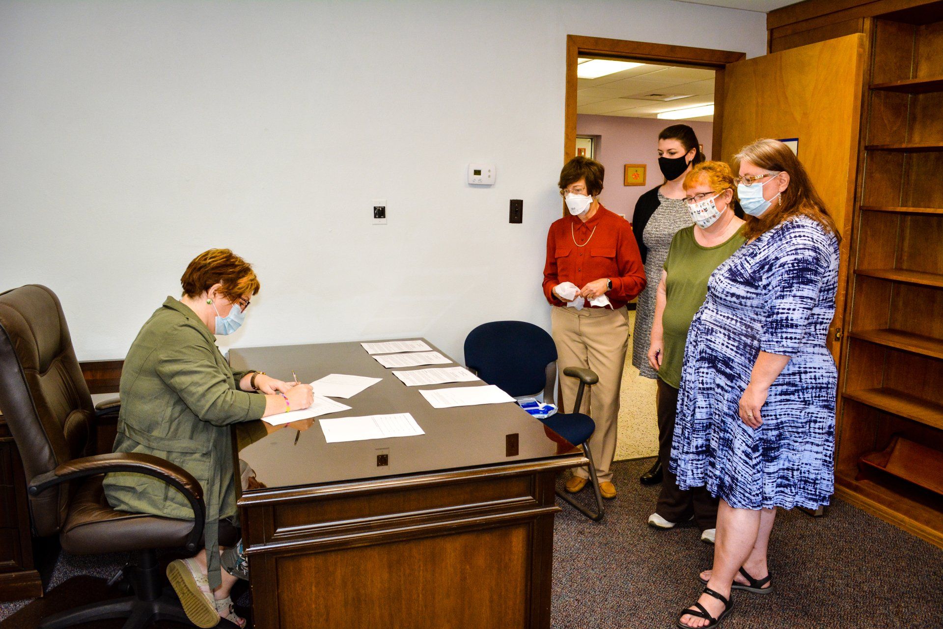 Office meeting with one person seated at a desk and three masked people standing nearby