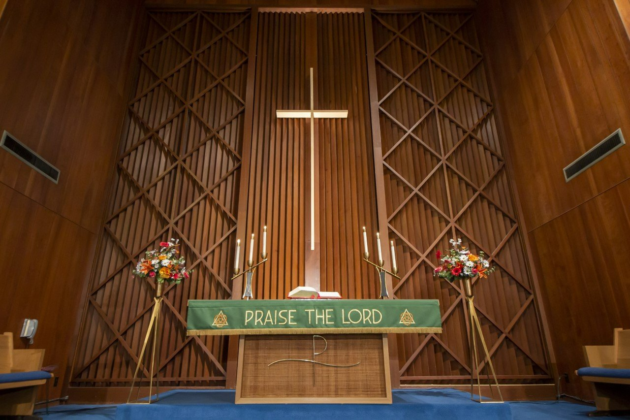 Church altar with wooden backdrop, cross, floral arrangements, and banner reading “PRAISE THE LORD”