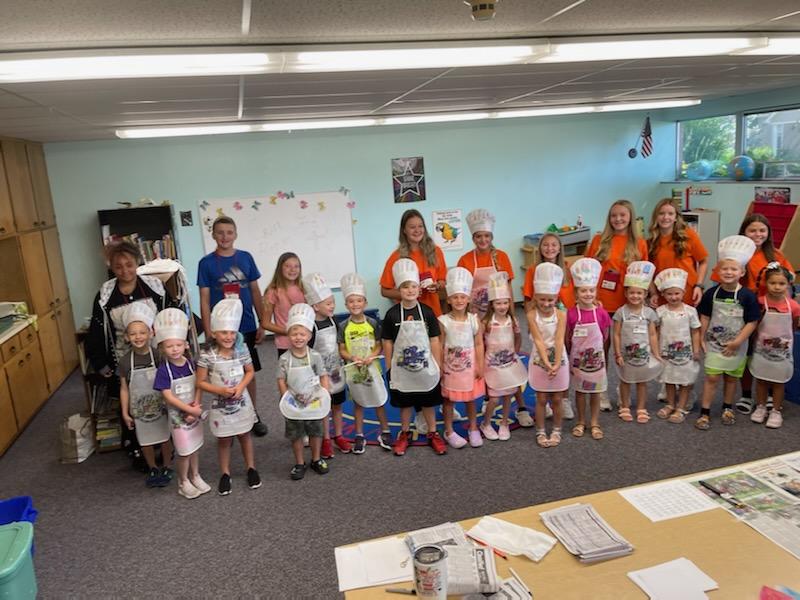 Group of children in aprons and chef hats posing in a classroom, holding signs and smiling.
