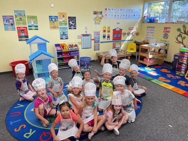 Children sitting on a blue rug in a colorful classroom, many wearing paper hats and smiling at the camera.