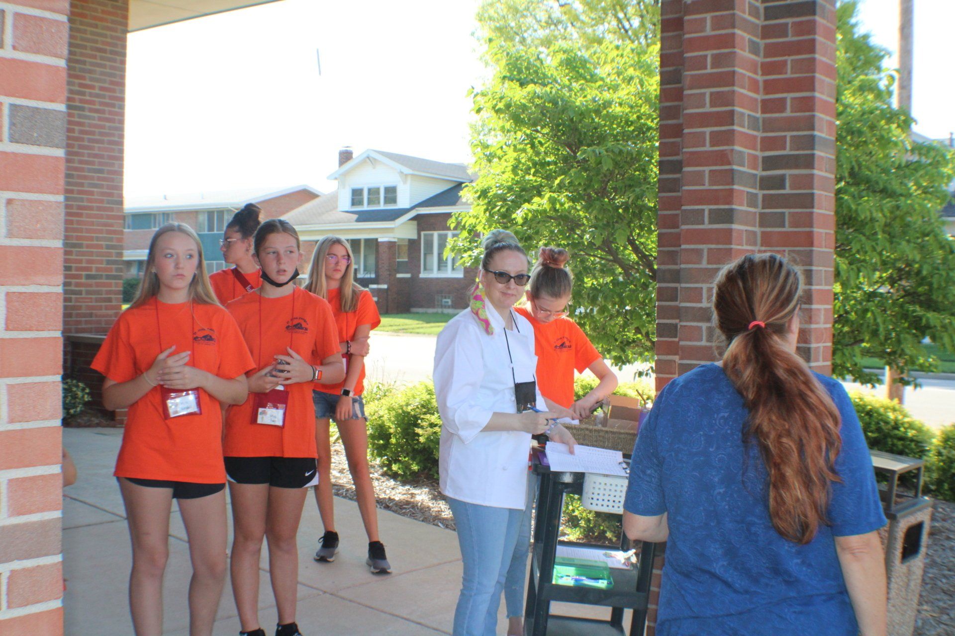 Volunteers in orange shirts serving food to a woman at a brick walkway outdoors