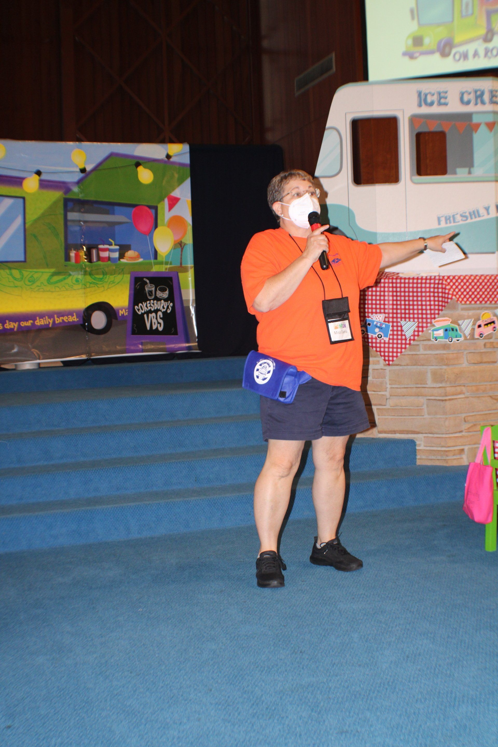 Person in an orange shirt speaking into a microphone and pointing inside an ice cream shop.
