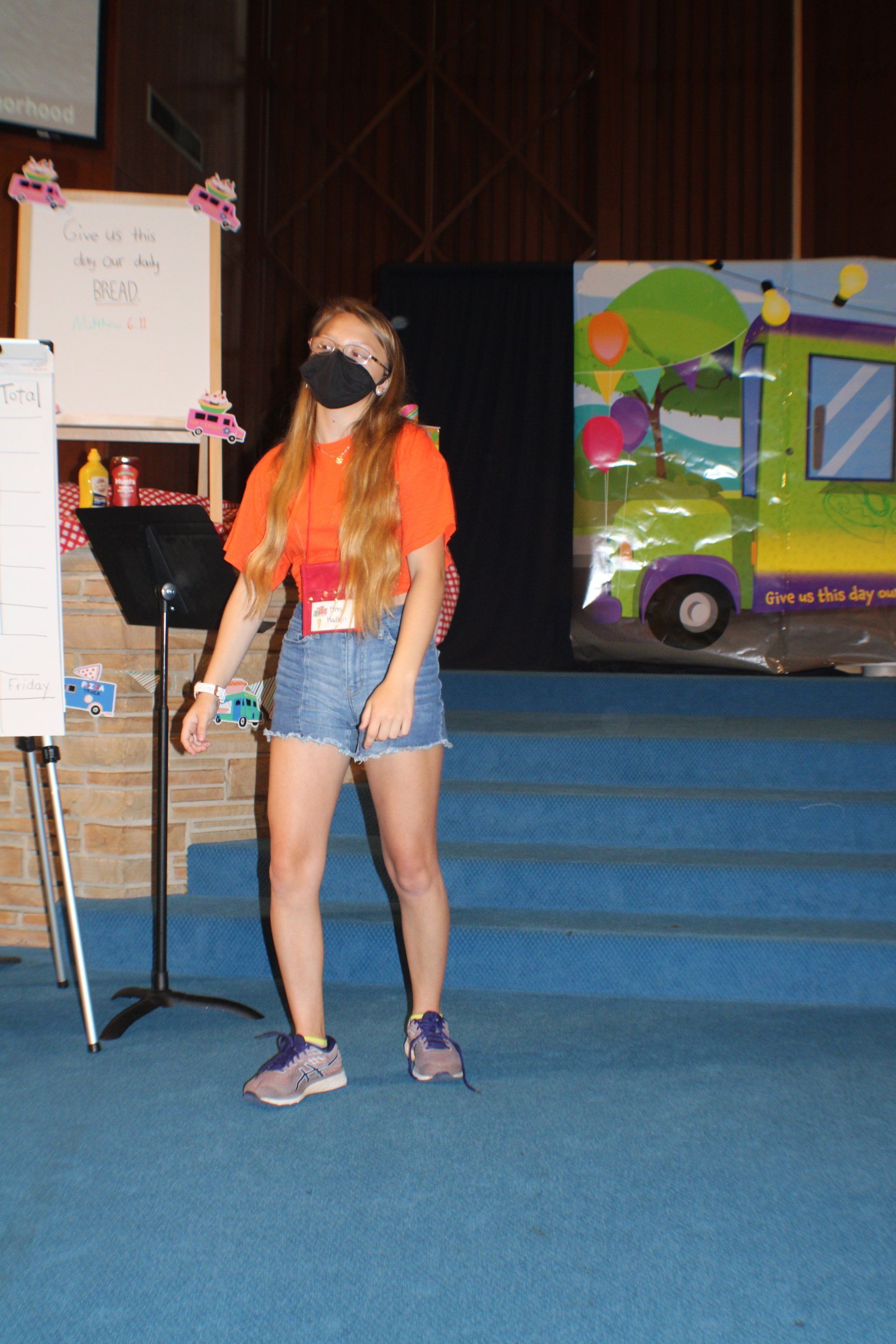 Masked woman in orange shirt and denim shorts standing on a blue carpeted stage beside an easel