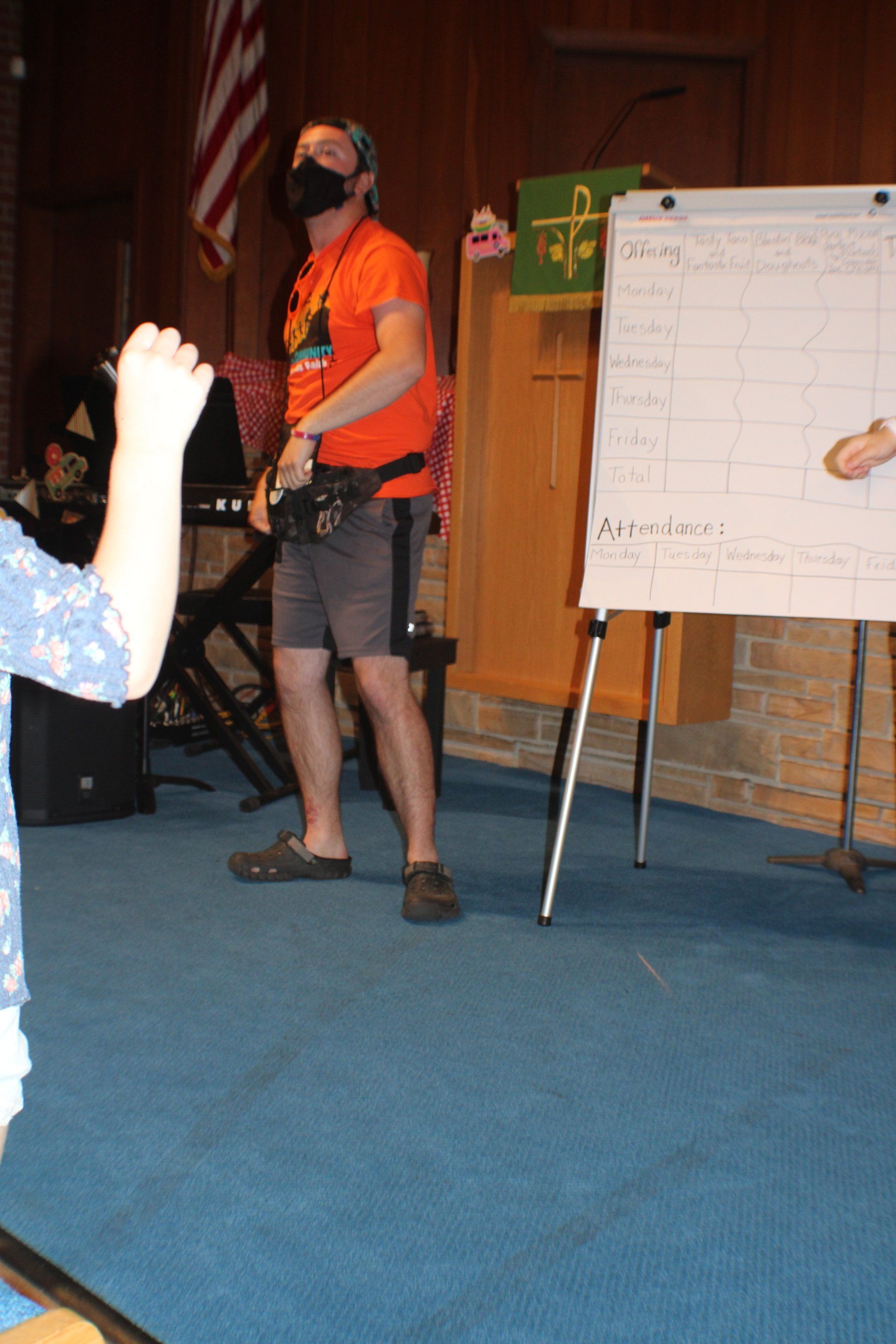 Man in orange shirt and shorts speaking beside a whiteboard in a church hall