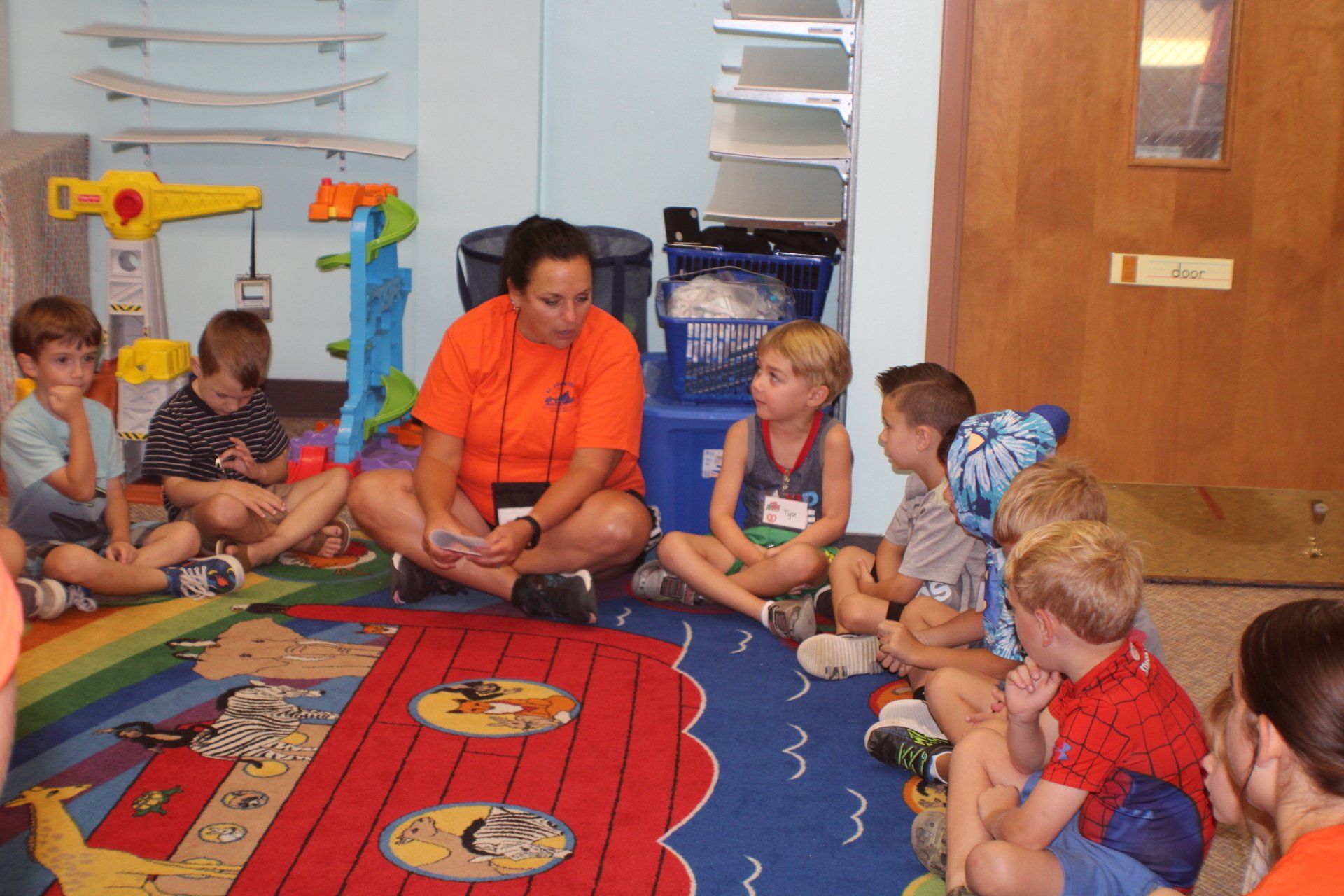 Children sit in a circle with a teacher on a colorful classroom rug.