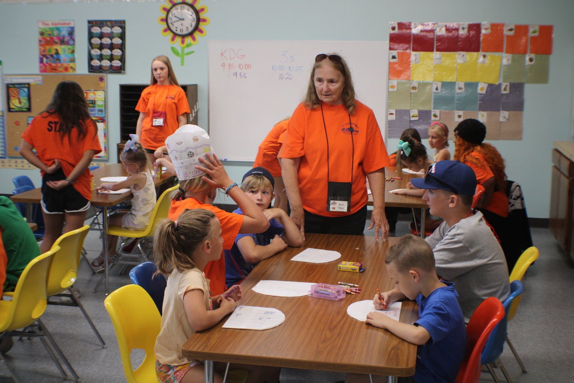 Children and adults in orange shirts gather around a table in a classroom for a group activity.