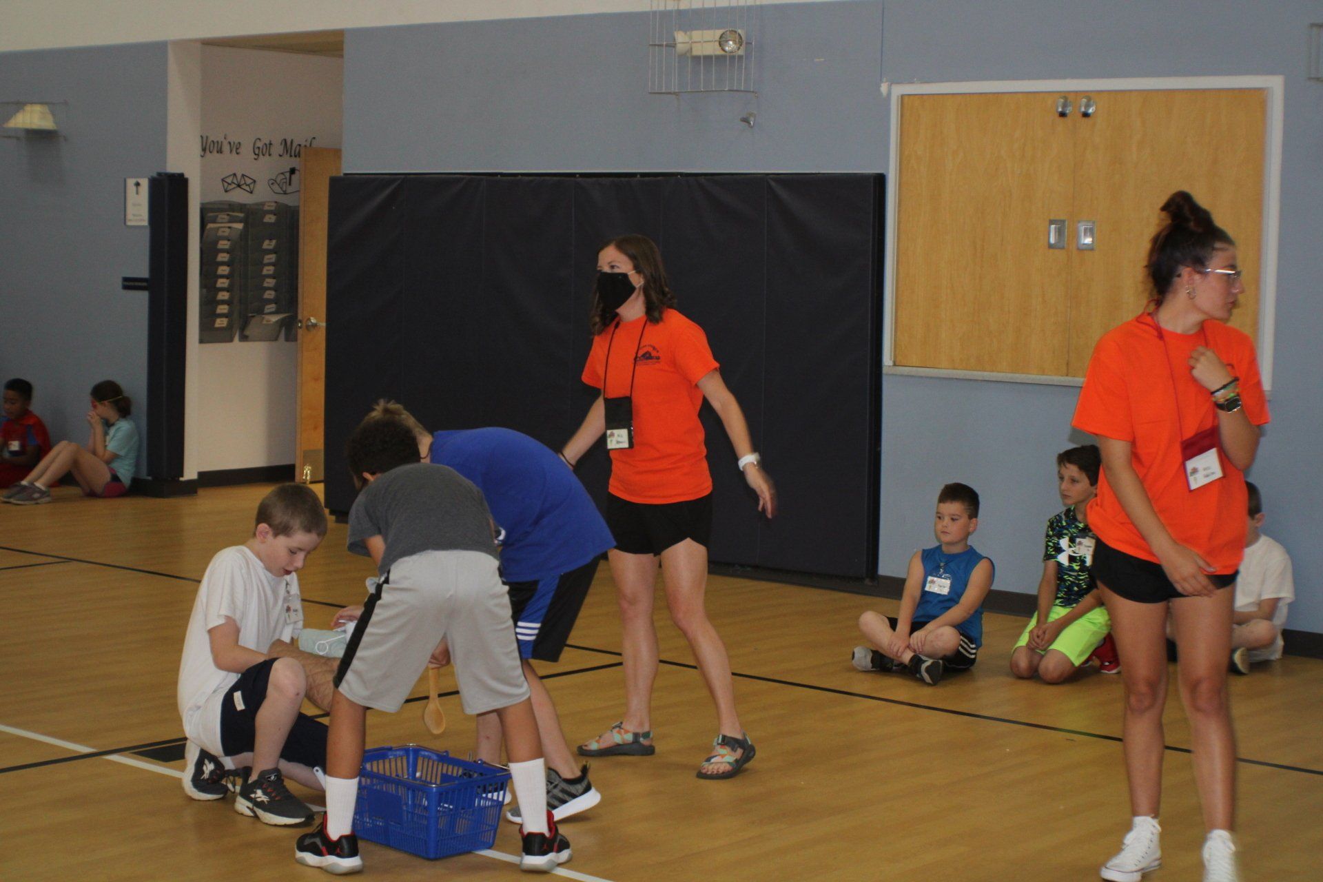 People in a gym play a seated game while others in orange shirts stand nearby