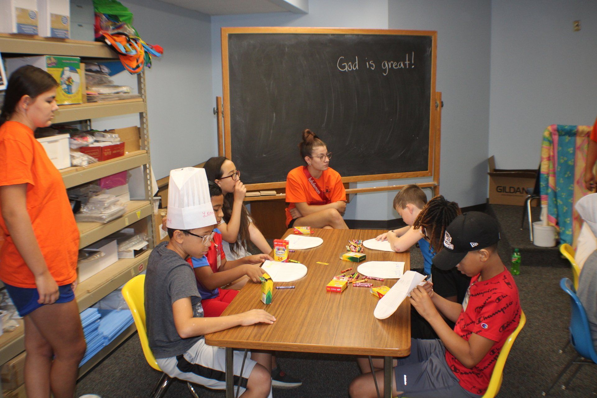 Children seated at a classroom table with a teacher, doing an activity near a chalkboard